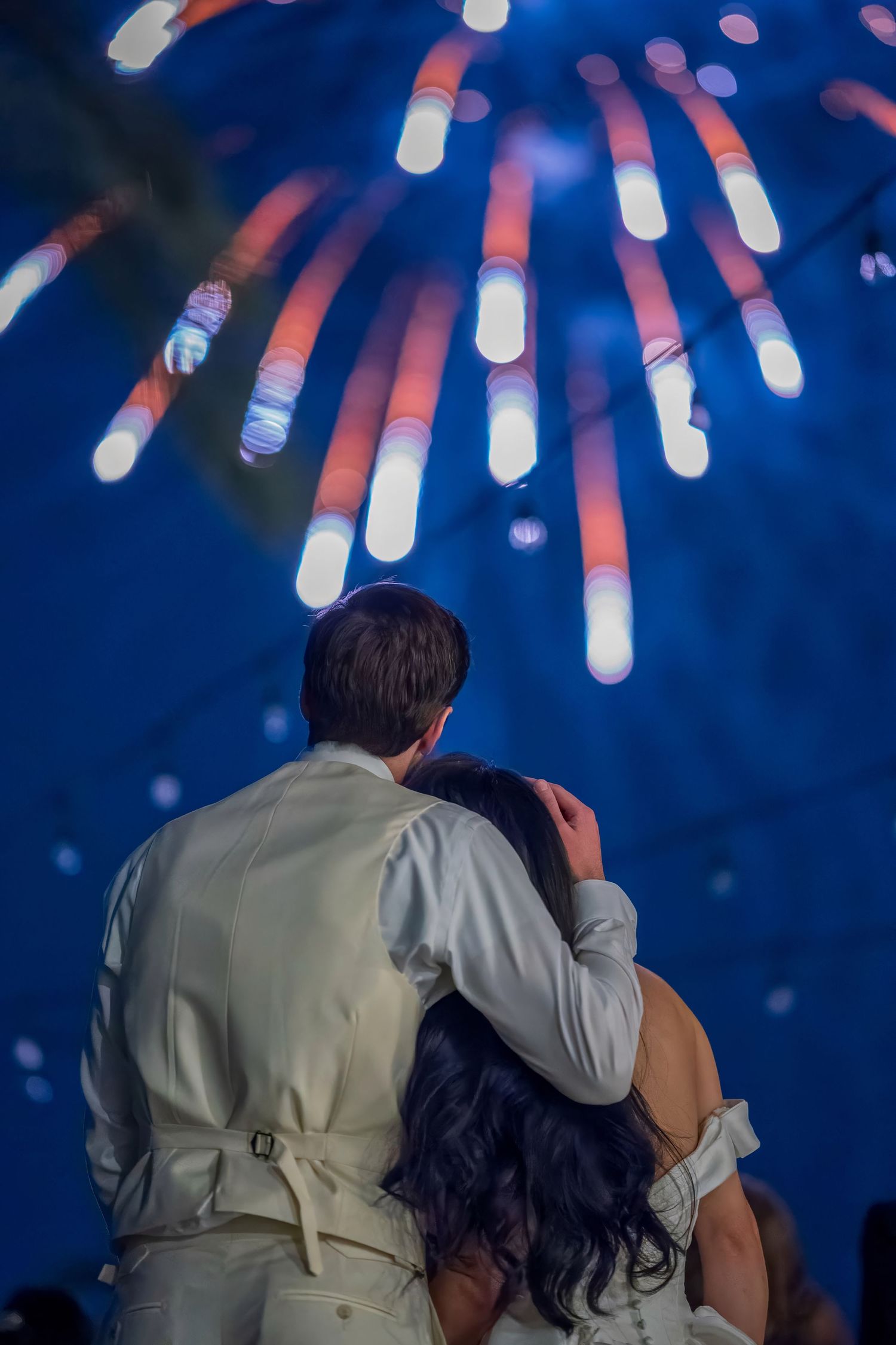 Couple embraces while watching fireworks display against dark blue night sky.