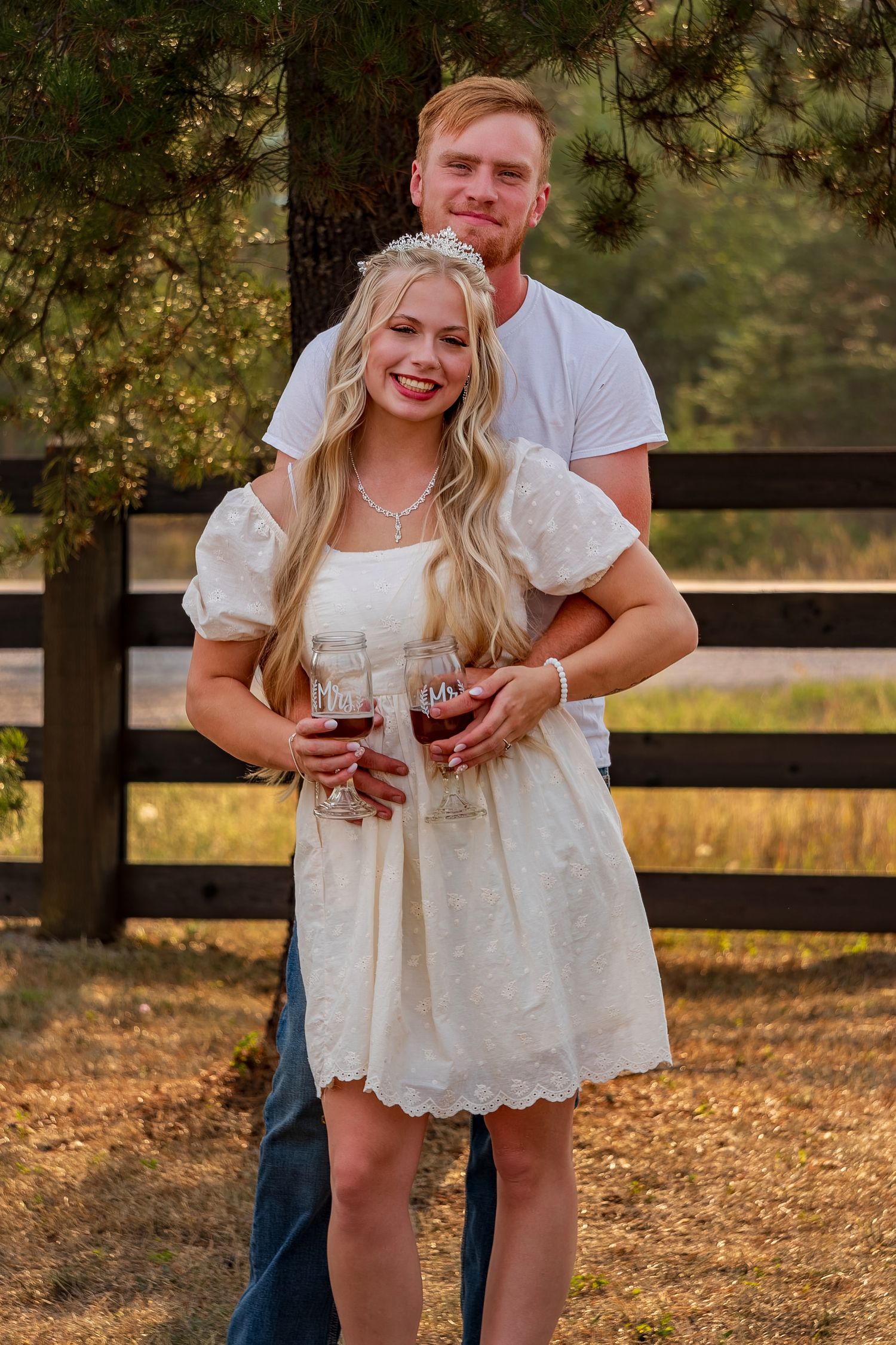 Couple embracing at sunset by rustic wooden fence holding wine glasses.