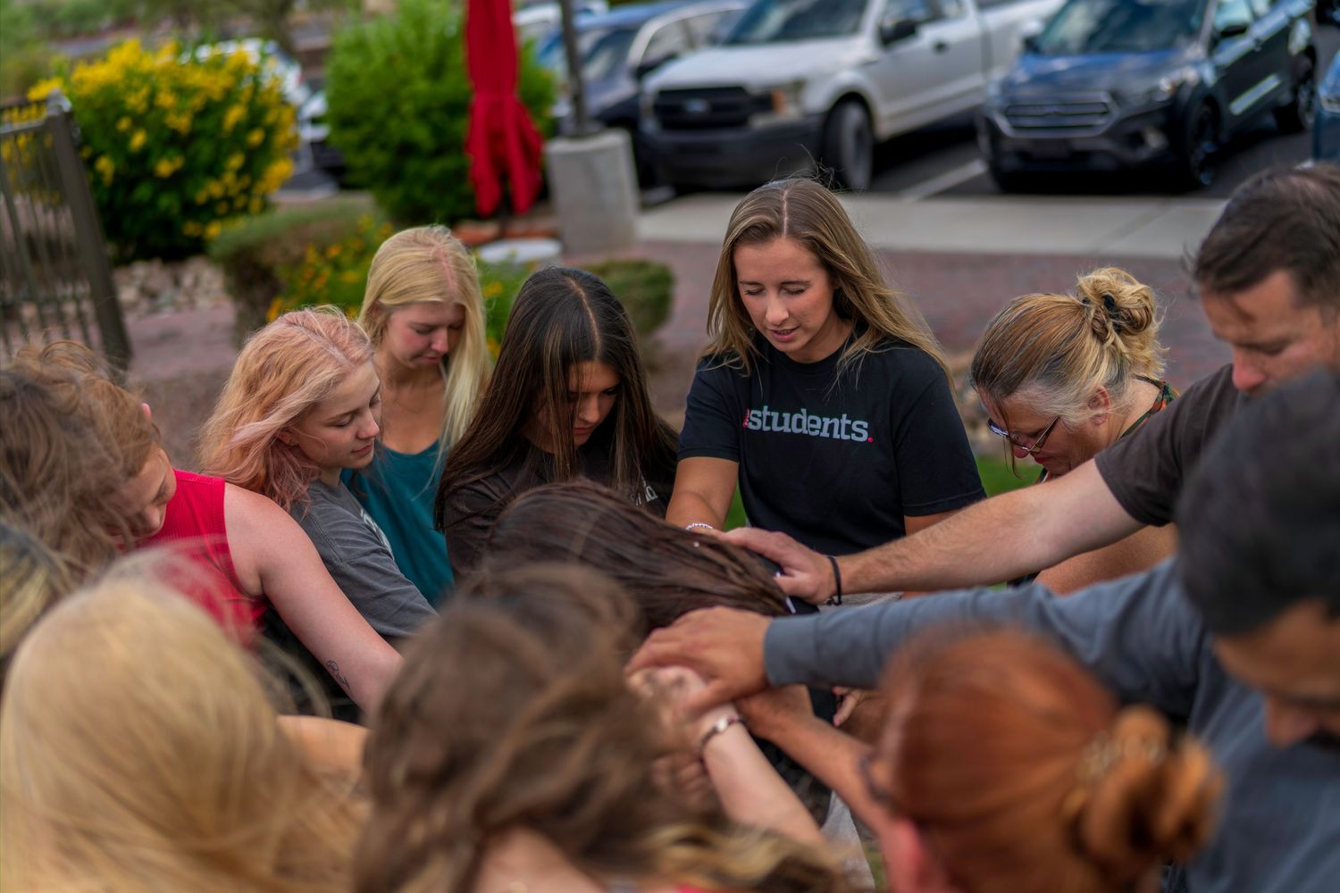 Group of young people praying together outdoors in a circle, bowing heads in a faith-based community gathering.