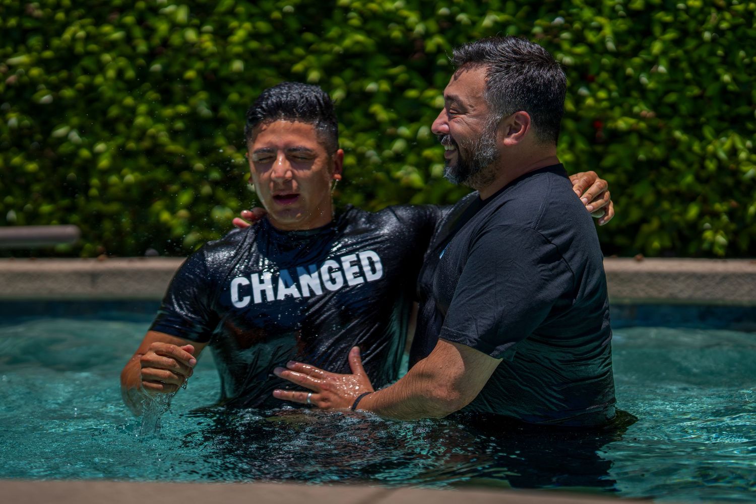 Two men standing in a pool during a baptism ceremony, one wearing a shirt that reads CHANGED.