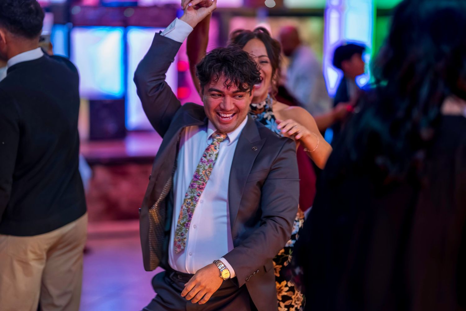 Young man in suit dancing energetically at a party, wearing a colorful tie, with guests in background.