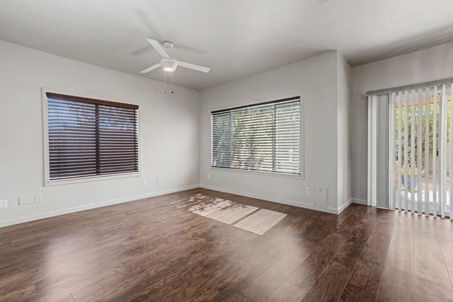 Bright empty living room with dark hardwood floors, white walls, ceiling fan, and large windows with blinds.