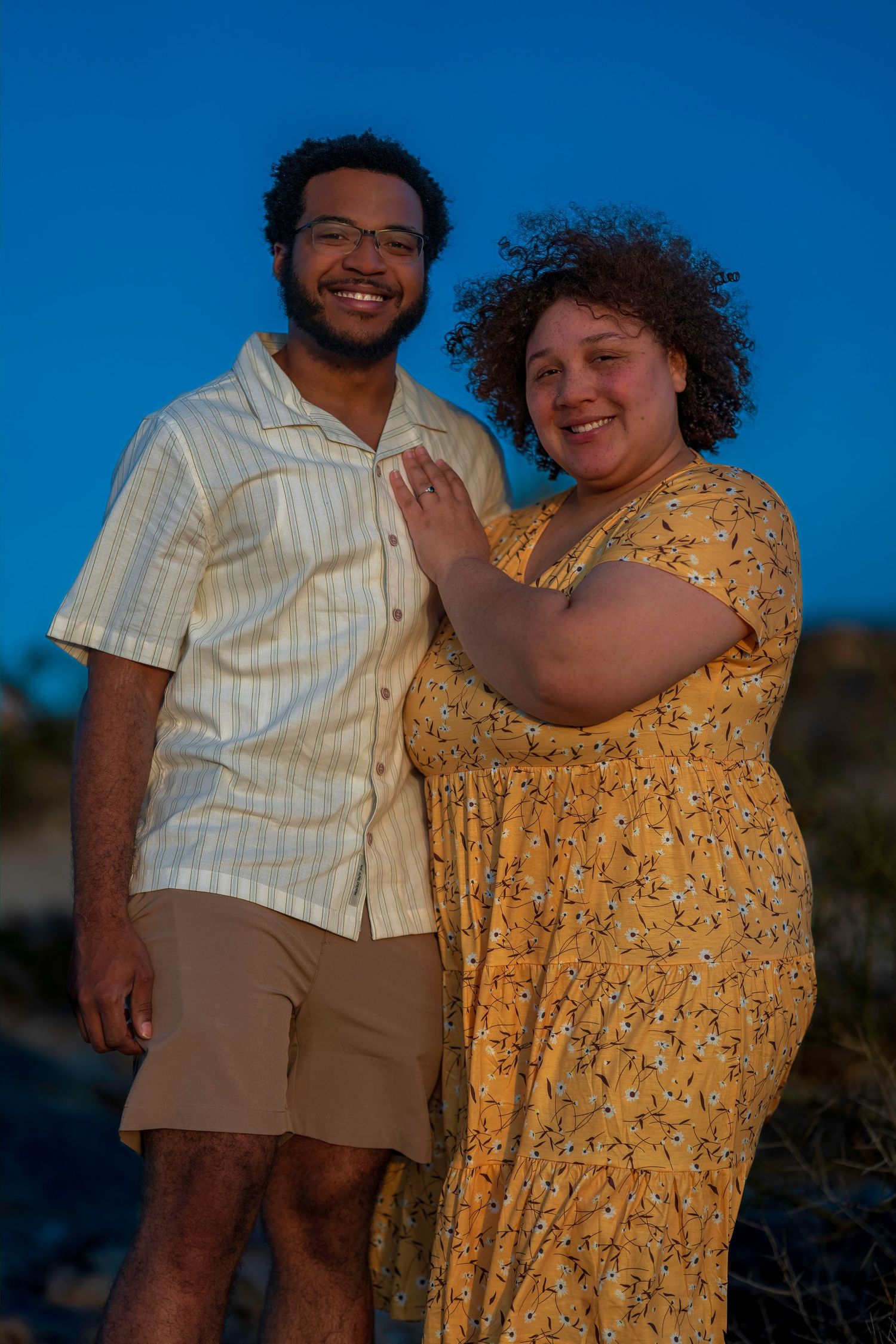 Smiling couple poses outdoors at dusk, man in striped shirt and woman in yellow floral dress against blue sky.