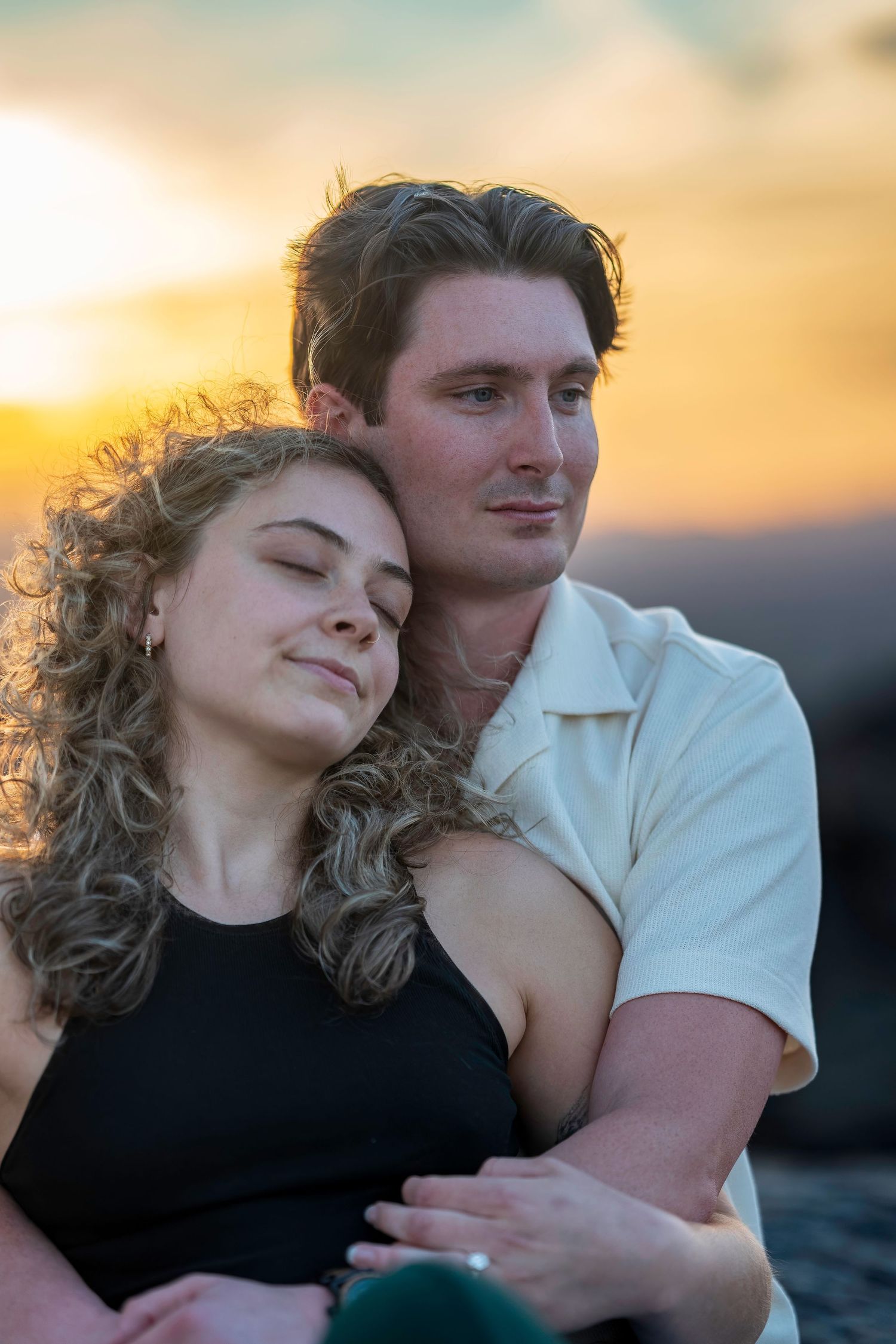Couple embracing at sunset, woman resting her head on mans shoulder with colorful sky behind.