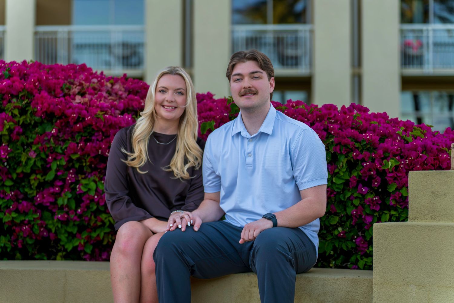 A young couple smiles while sitting together in front of vibrant pink bougainvillea flowers outdoors.