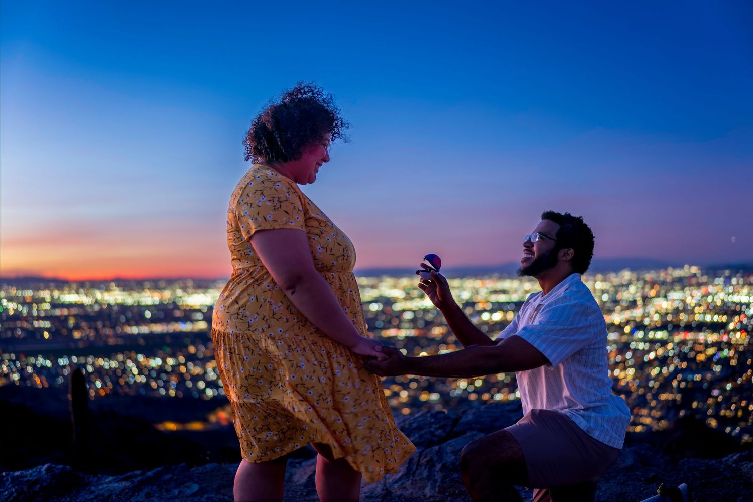 Man proposes on one knee to woman in yellow dress against stunning city lights at night on hilltop.