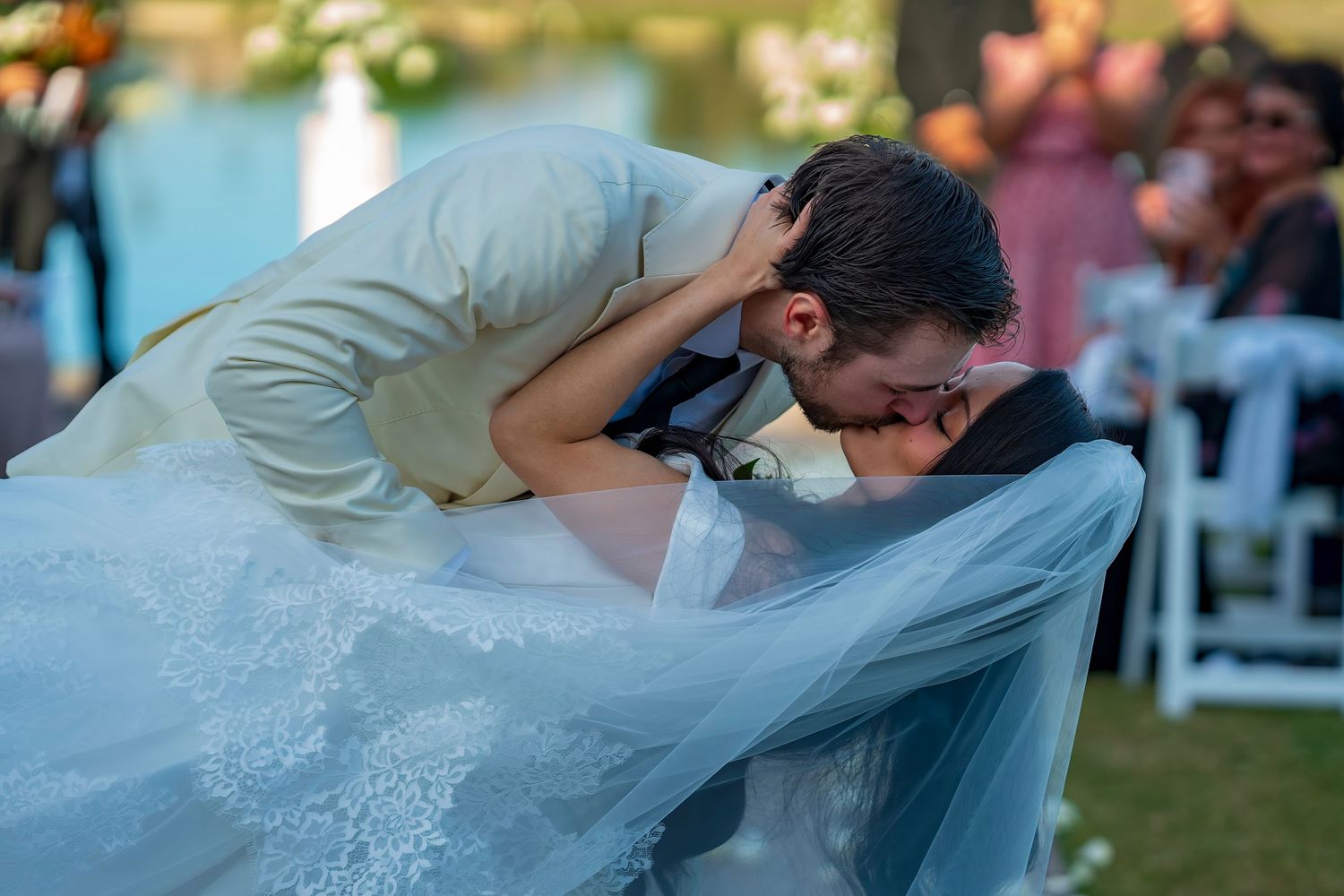 A wedding couple shares a tender moment under a flowing veil against a vibrant blue sky.