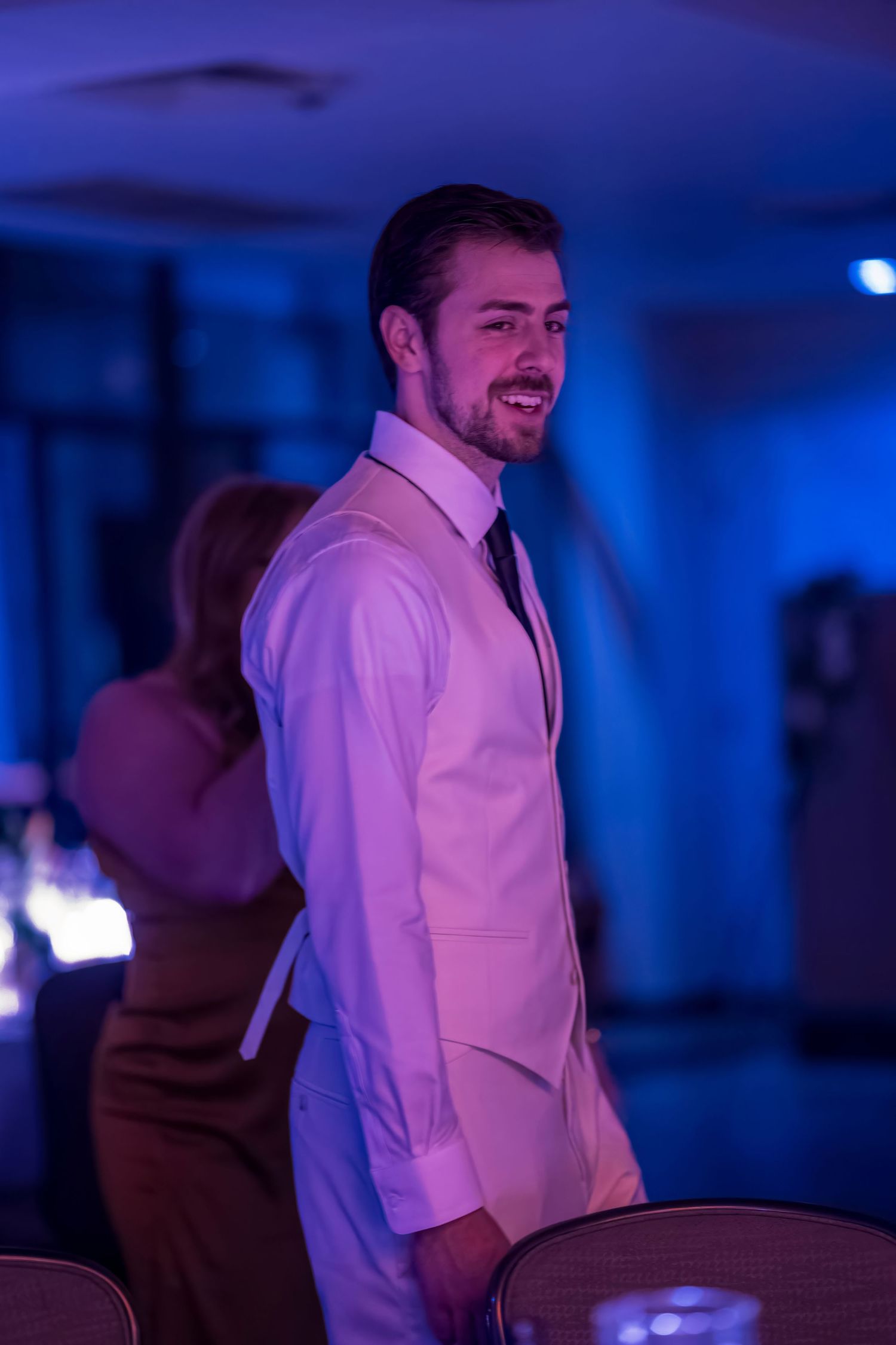 A person in formal attire smiles while standing in a room with blue ambient lighting.