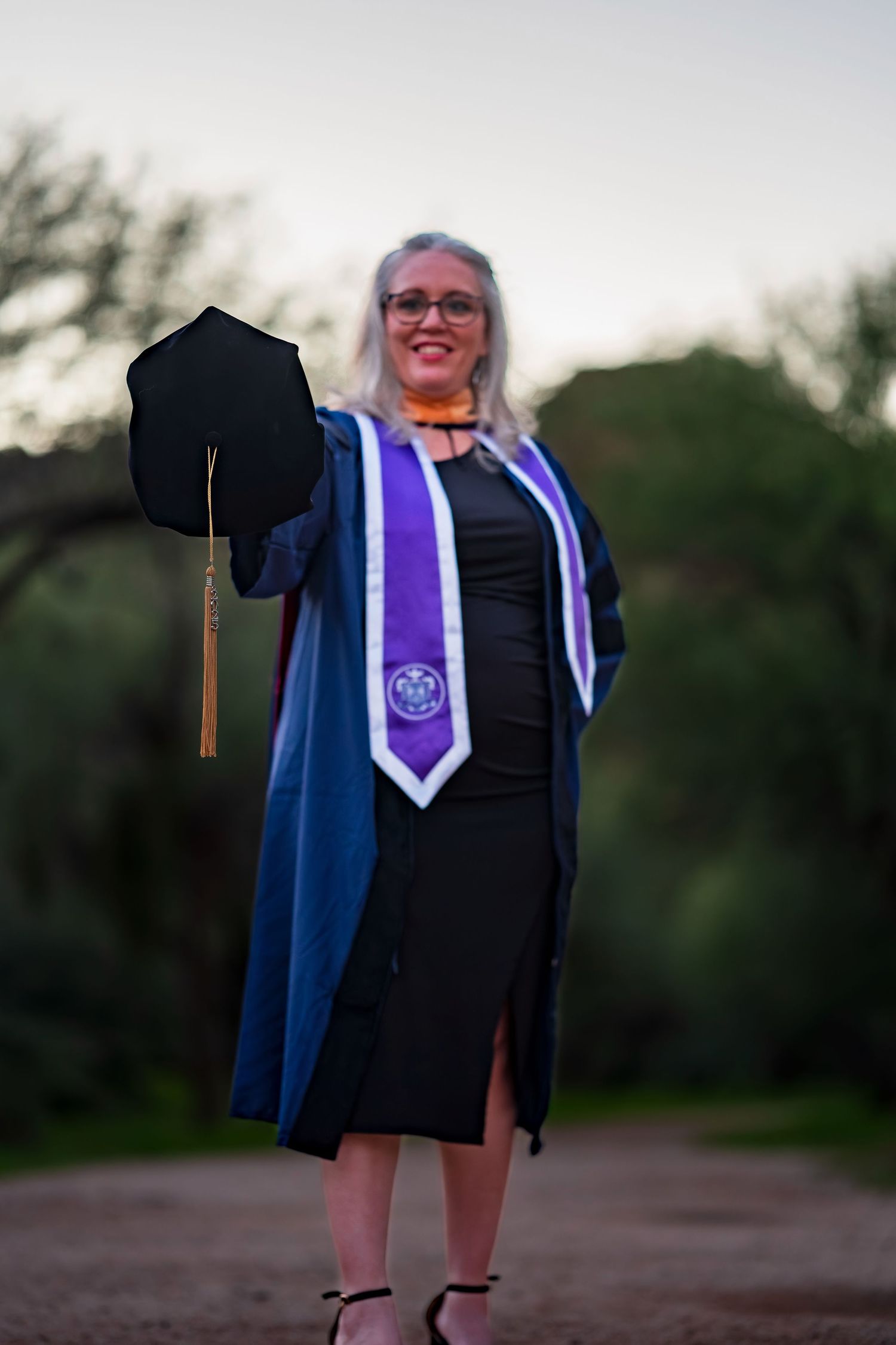 Graduate holding mortarboard cap with tassel while wearing blue and purple academic regalia.