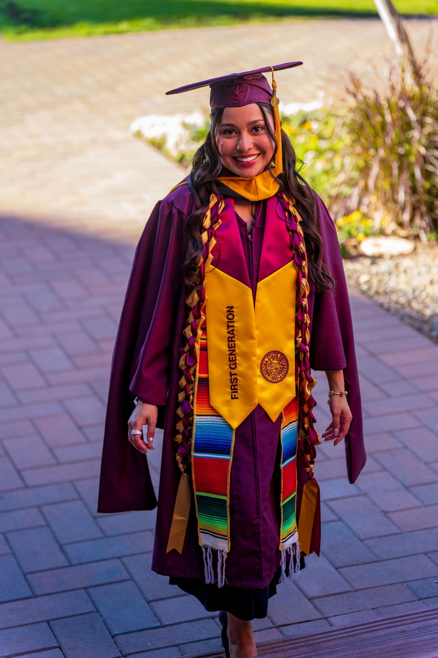 Graduate wearing maroon cap and gown with yellow stole and colorful graduation stole outdoors.