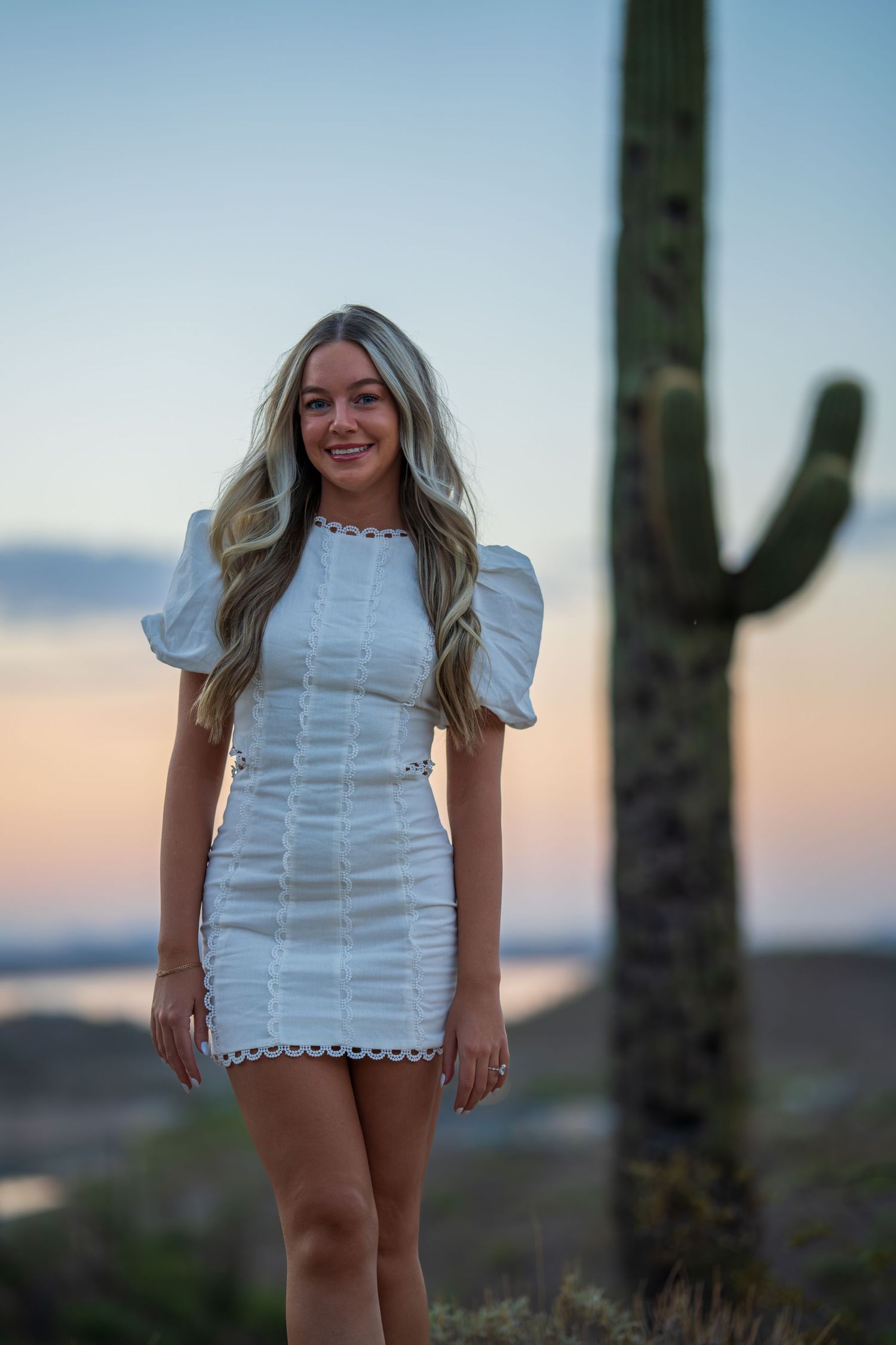 Person in white lace dress poses in desert landscape with saguaro cactus at sunset.