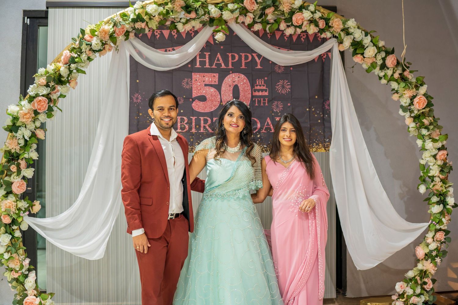 Three people stand under decorated arch at 50th birthday celebration wearing formal attire.