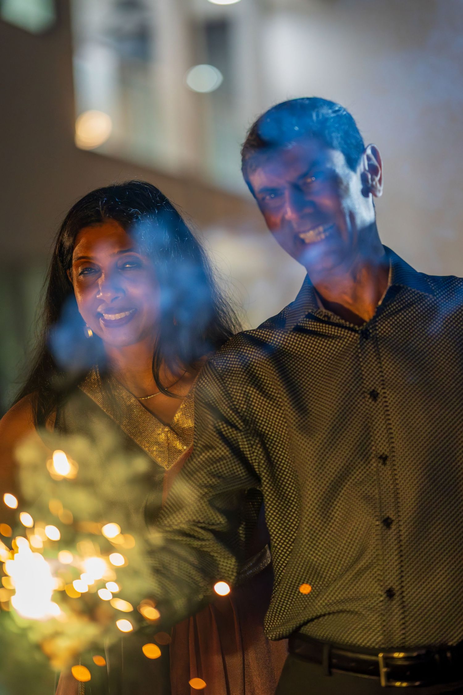 A couple smiling while holding sparklers with bokeh lights in the background.