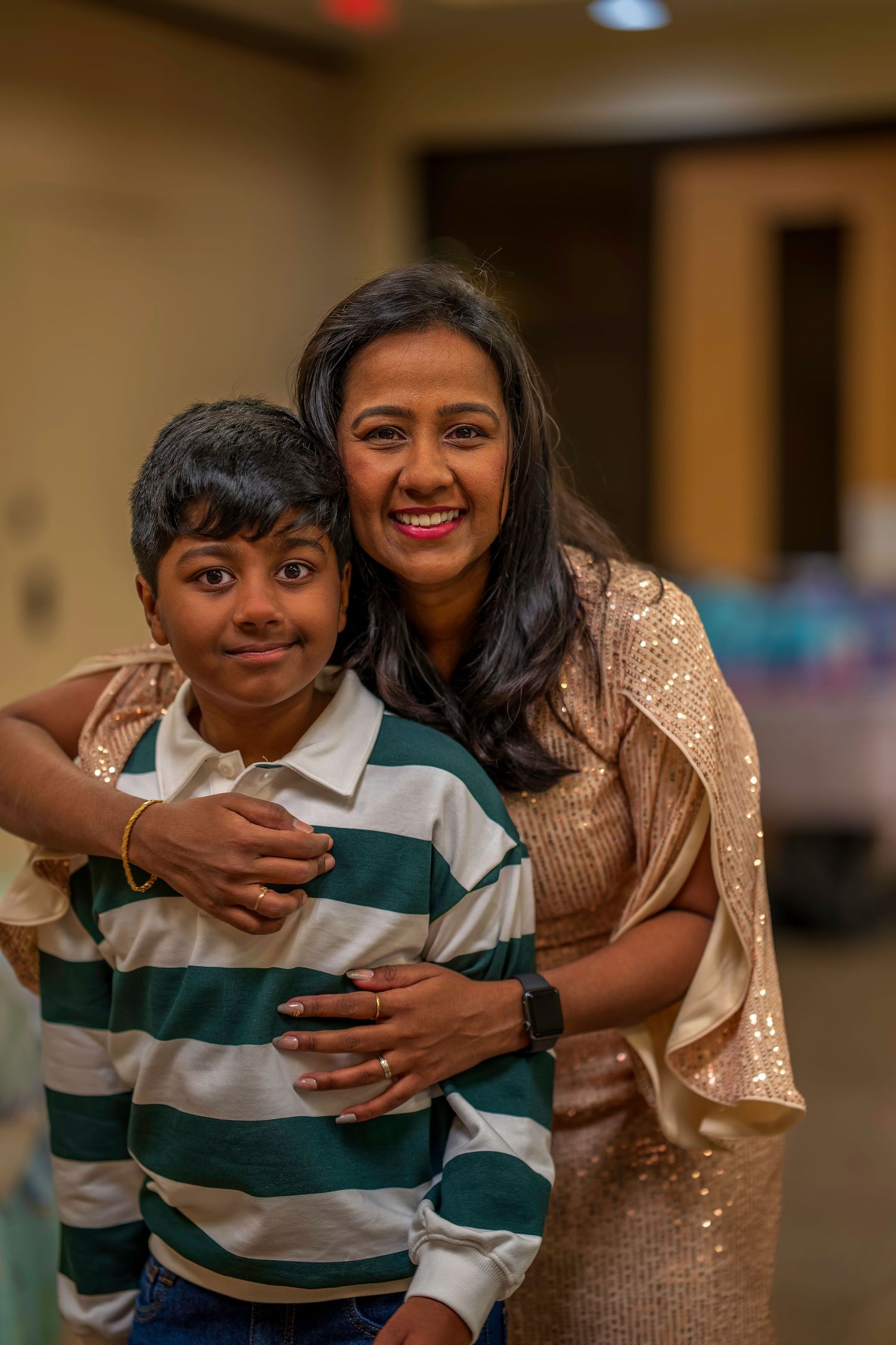 A woman in a gold sequin dress smiles while hugging a young boy in a striped shirt at an indoor event.