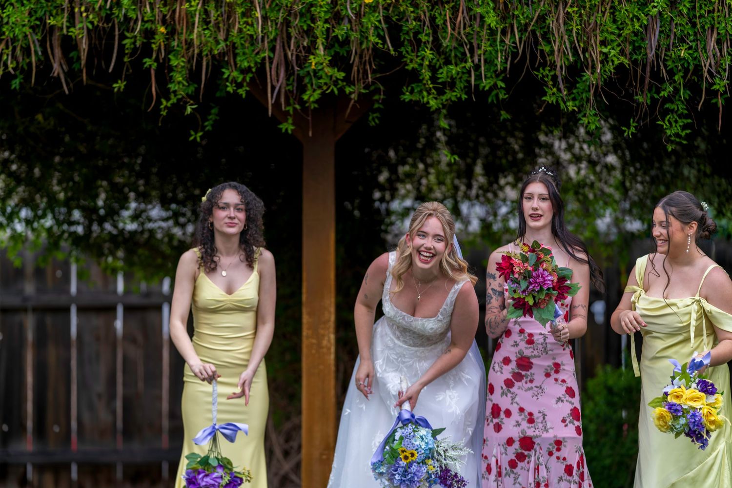 Bride in white gown laughs with three bridesmaids in yellow and floral dresses holding colorful bouquets outdoors.