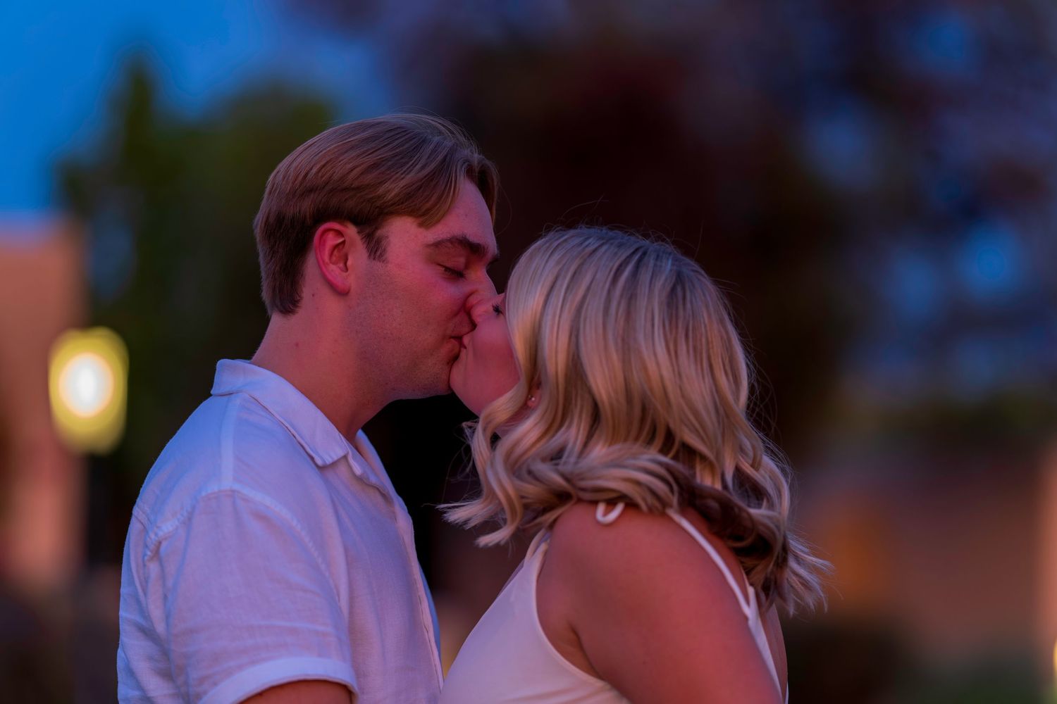 A newly engaged couple shares a romantic kiss at dusk with a soft bokeh background.