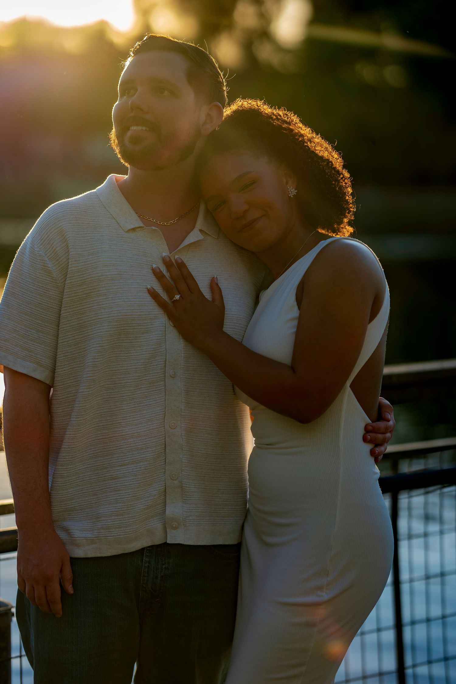 Couple embracing at golden hour sunset, woman in white dress leans on mans shoulder showing engagement ring.