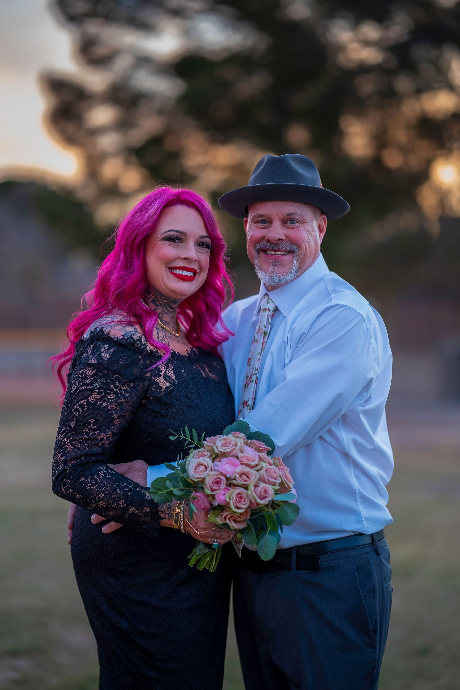 Couple posing at sunset, woman with pink hair in black lace dress holding pink roses, man in white shirt and gray fedora.