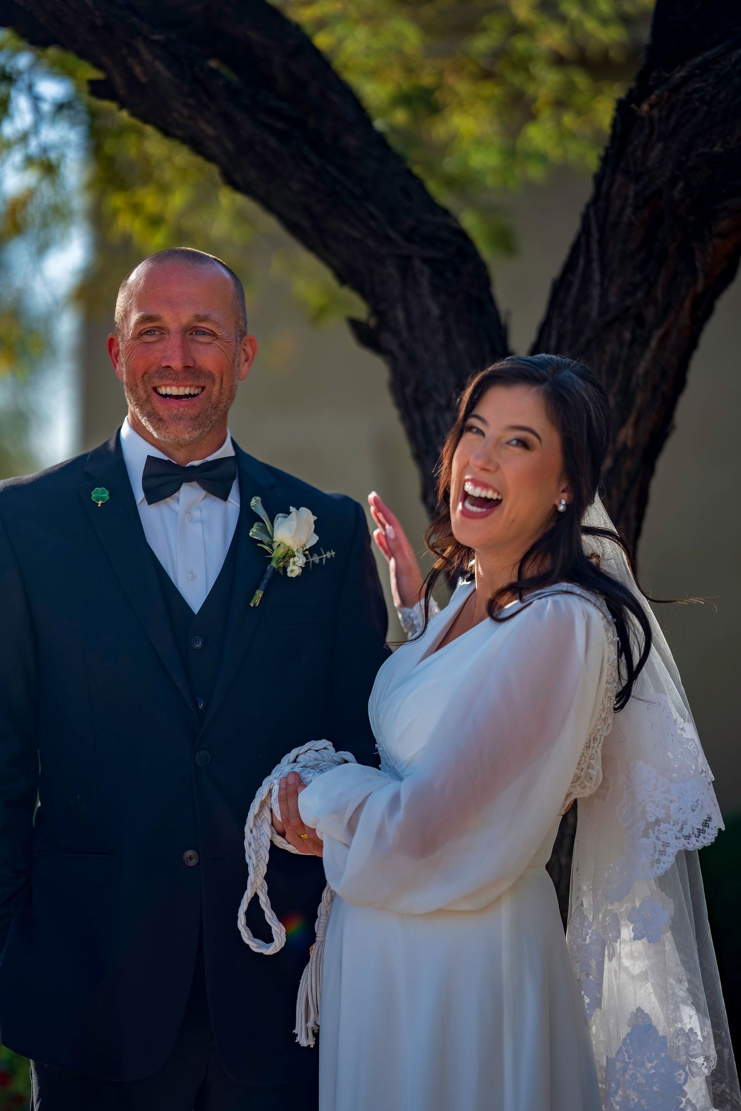 Laughing bride and groom pose together outdoors after wedding ceremony under tree.