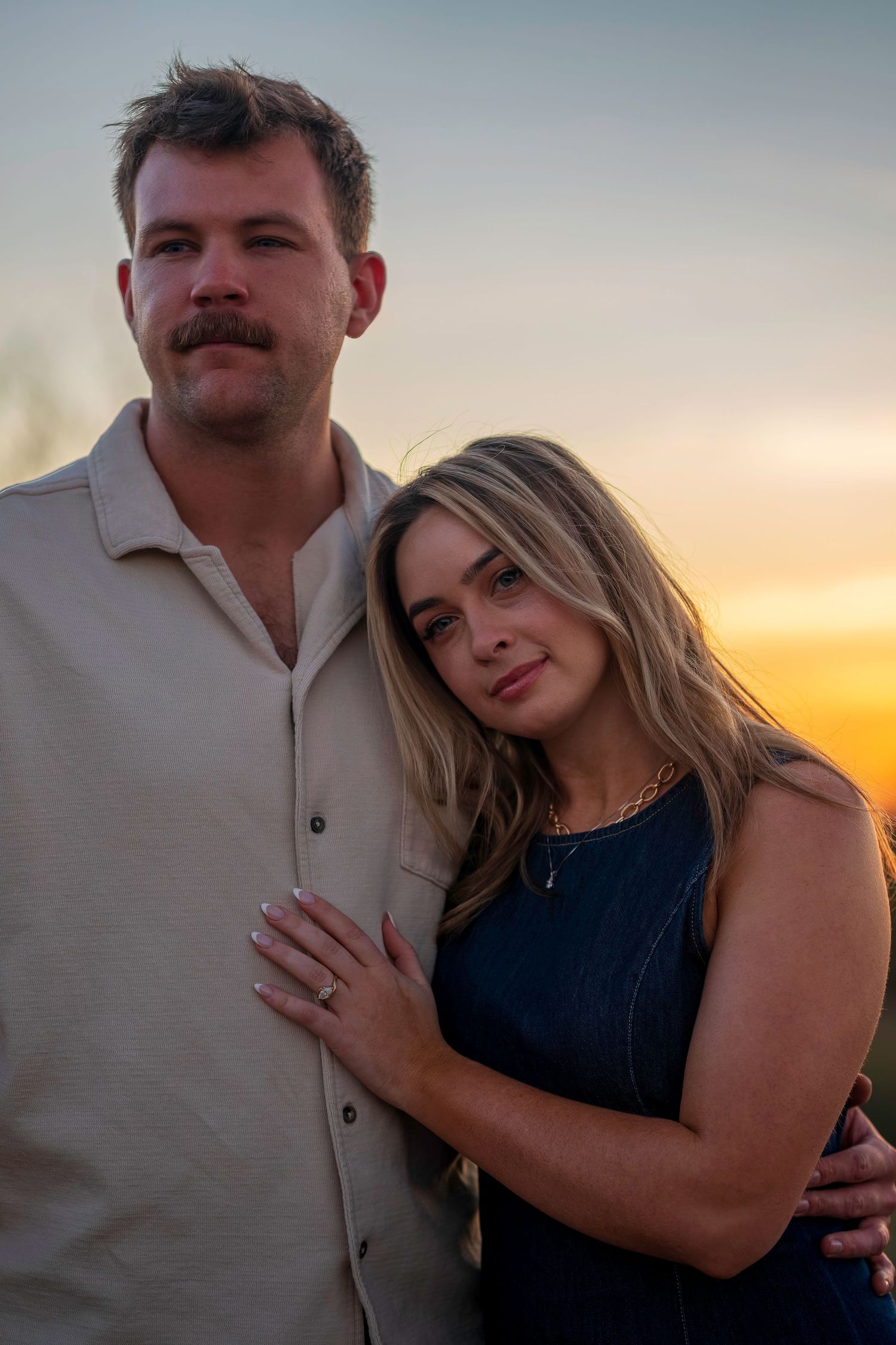 Couple posing together at sunset, woman leaning on man's shoulder, engagement ring visible on her hand.