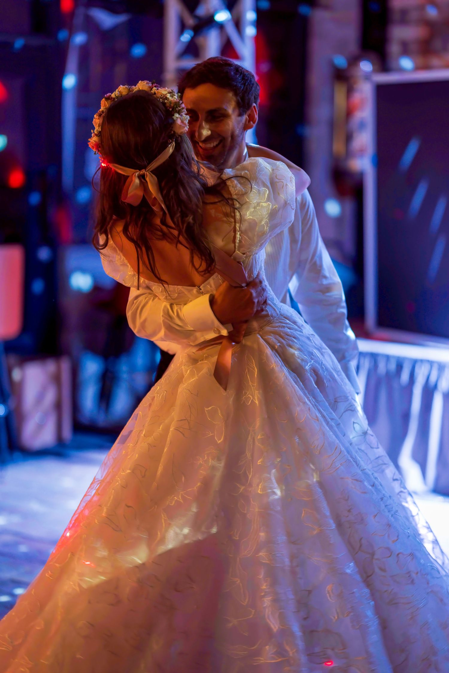Bride and groom share first dance, bride in elegant white ball gown with floral headpiece under colorful reception lights.