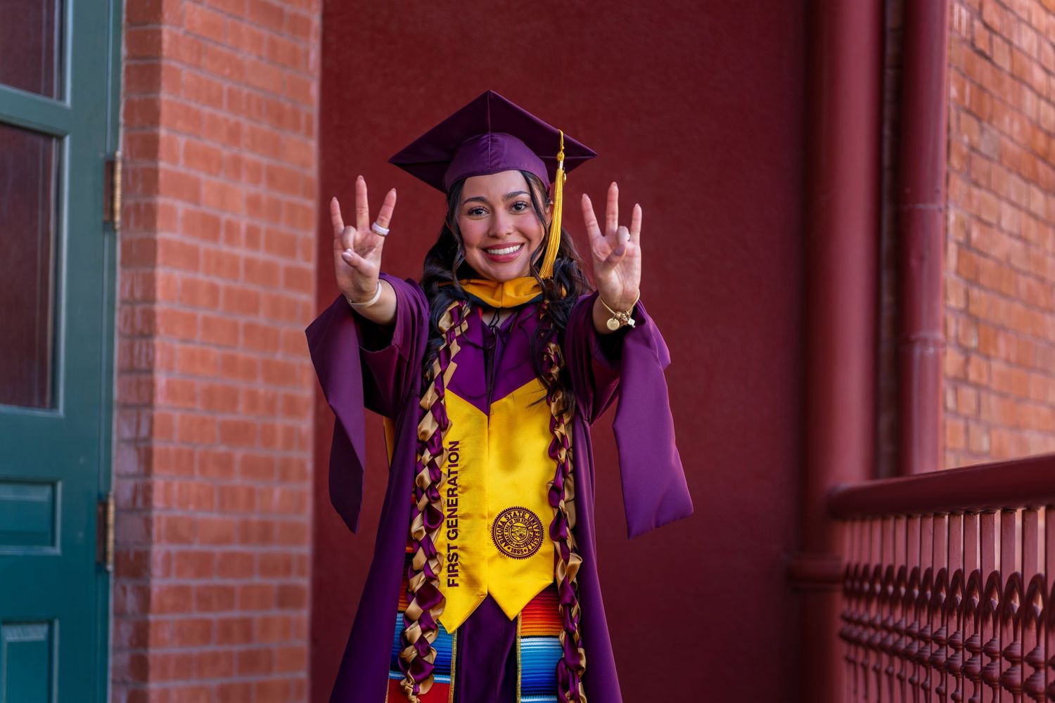 Graduate in purple graduation attire celebrates with enthusiastic gestures against brick building.
