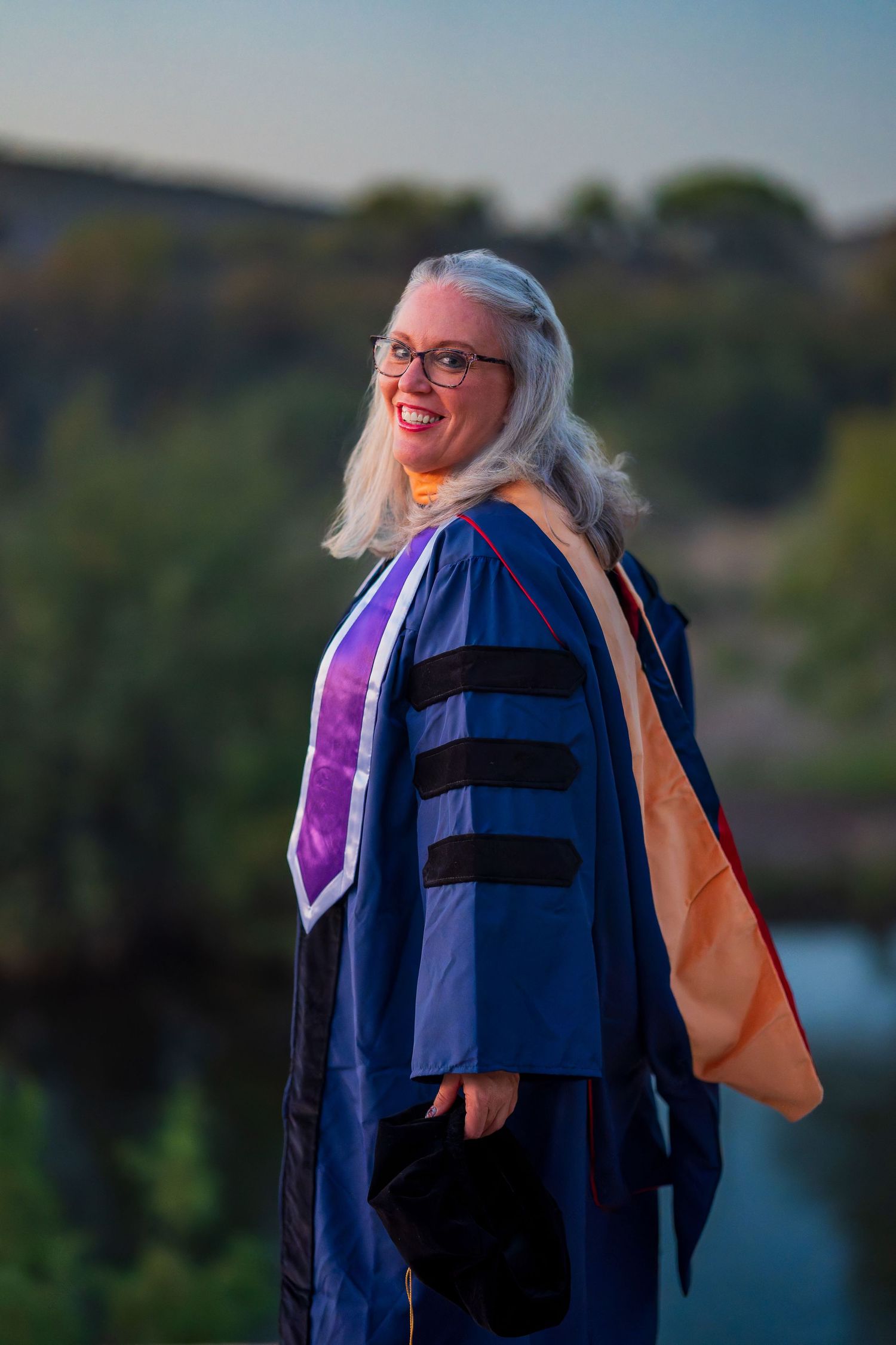 Graduate in blue doctoral regalia photographed against scenic natural landscape.