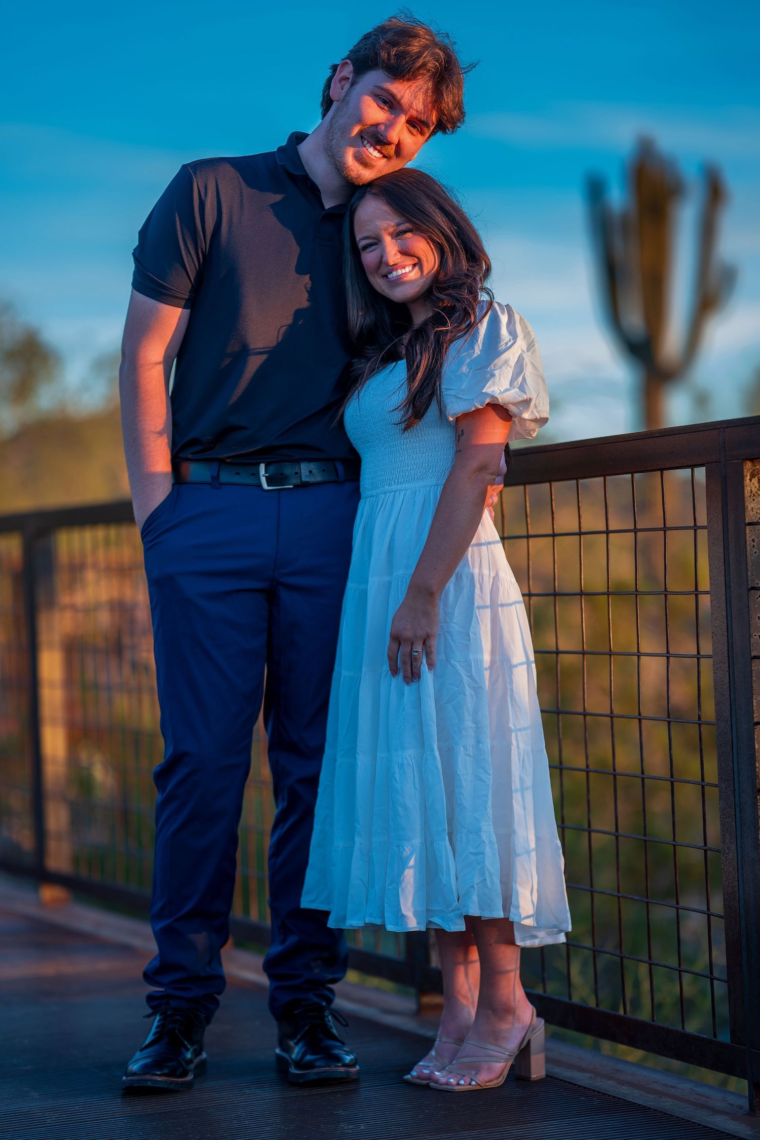 A couple stands together on a desert viewing deck during golden hour.