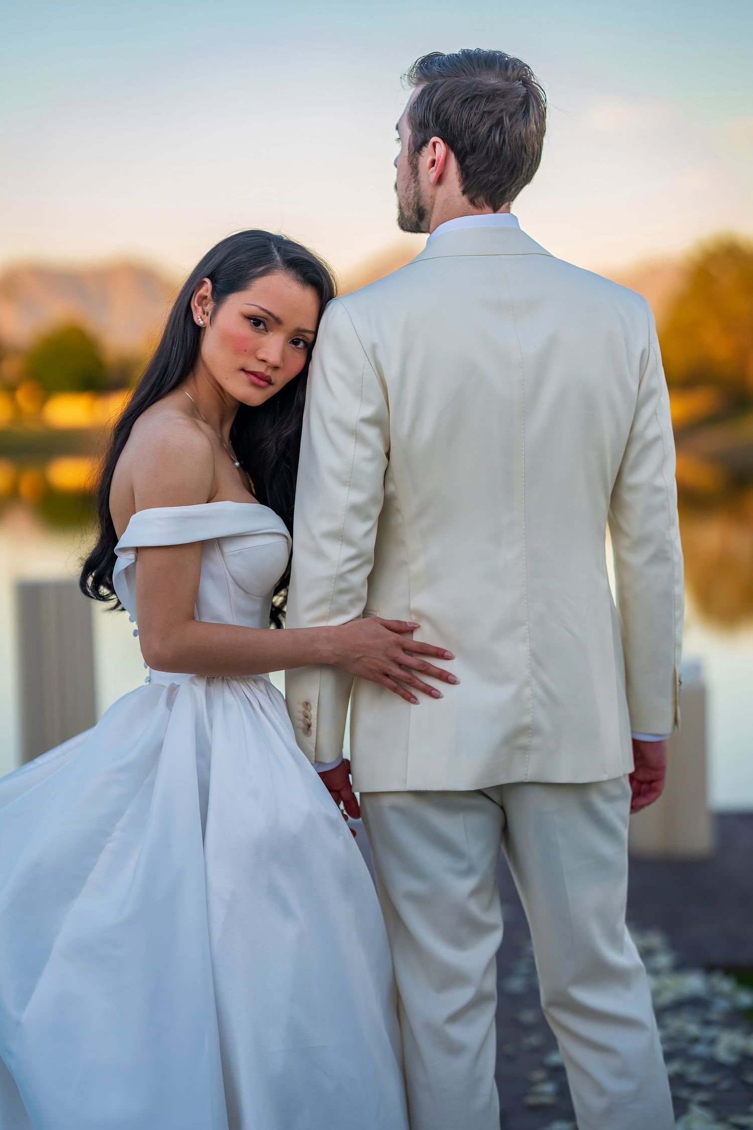 Couple in white wedding attire pose against sunset lake backdrop.