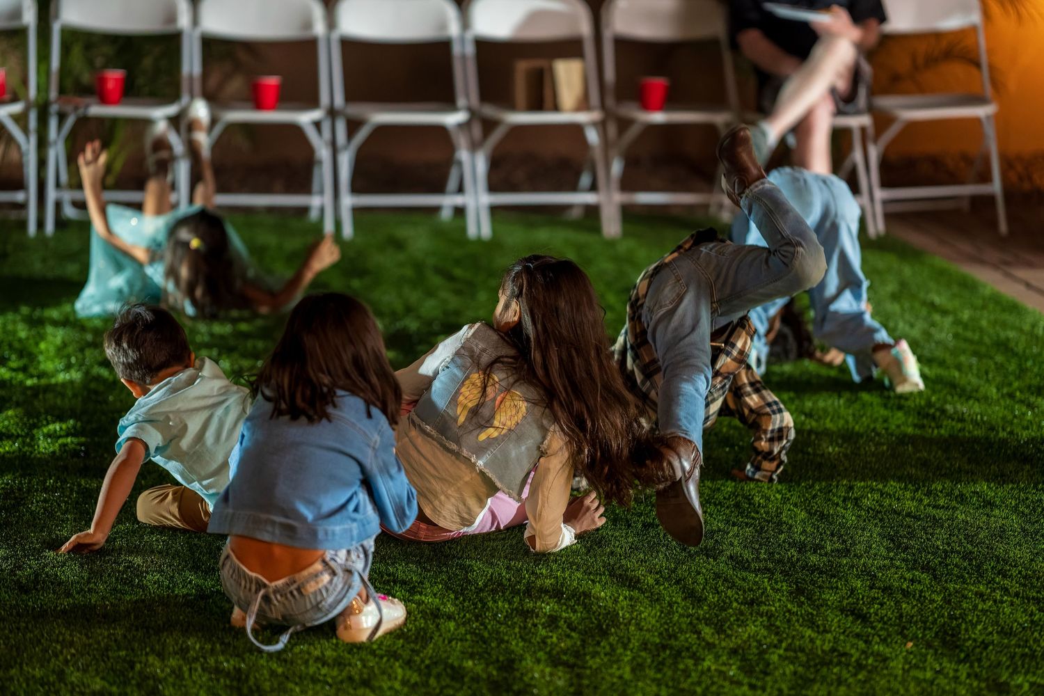 Group of people lying on grass near white folding chairs during an outdoor gathering.