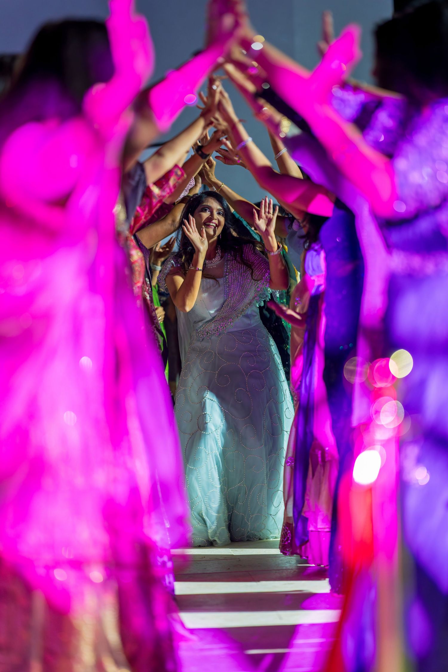 Dancers create tunnel with raised arms under vibrant pink and purple lighting.