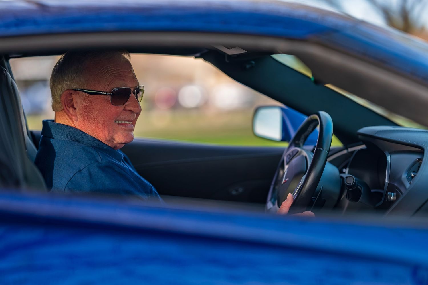 Elderly man wearing sunglasses smiling while sitting in the driver seat of a blue car.