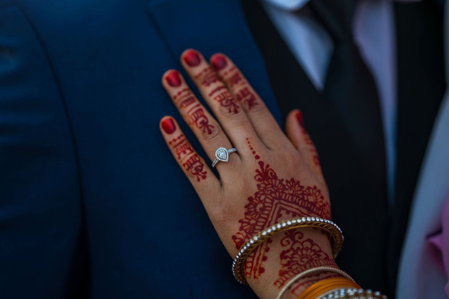 Close-up of a bride's hand with intricate red henna mehndi design and diamond engagement ring with gold bangles.