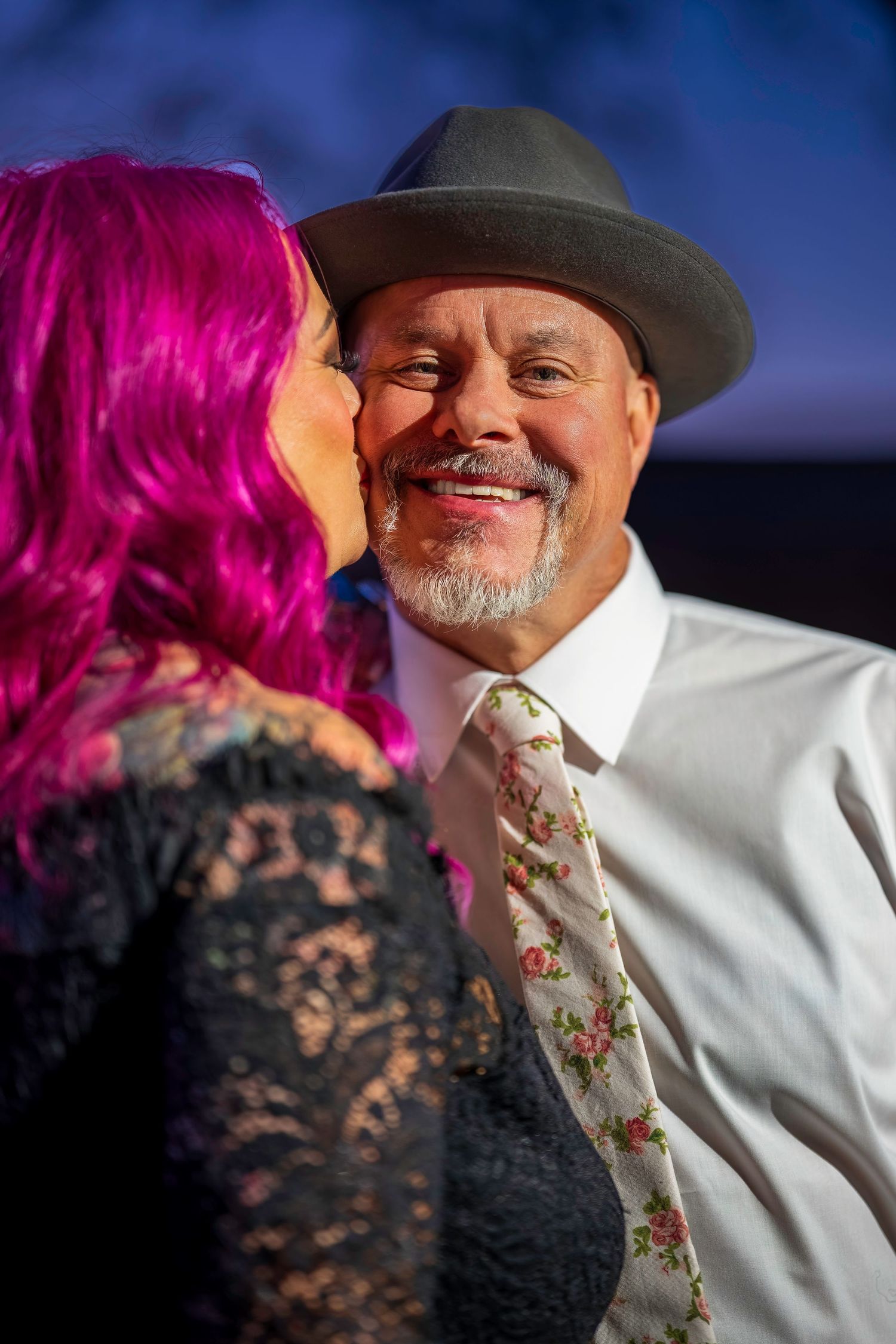 Woman with pink hair kissing smiling man on cheek, man wearing gray hat and floral tie at outdoor event.