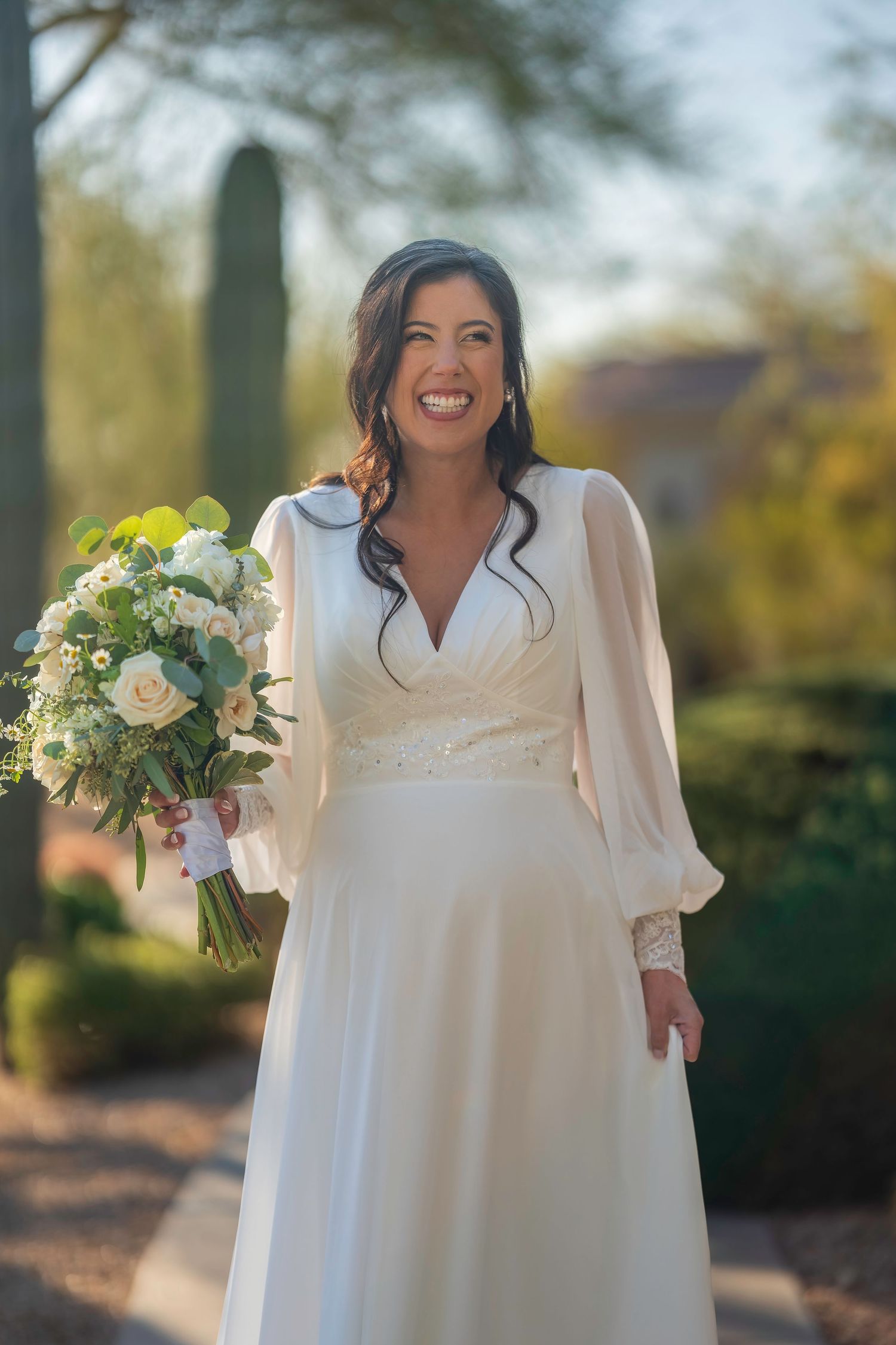 Smiling bride in elegant long-sleeve white wedding gown holding a bouquet of white and green flowers outdoors.
