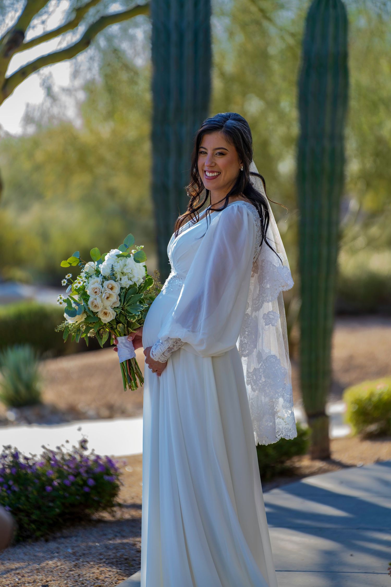 Pregnant bride in white long-sleeve gown with veil holding white floral bouquet in desert garden.