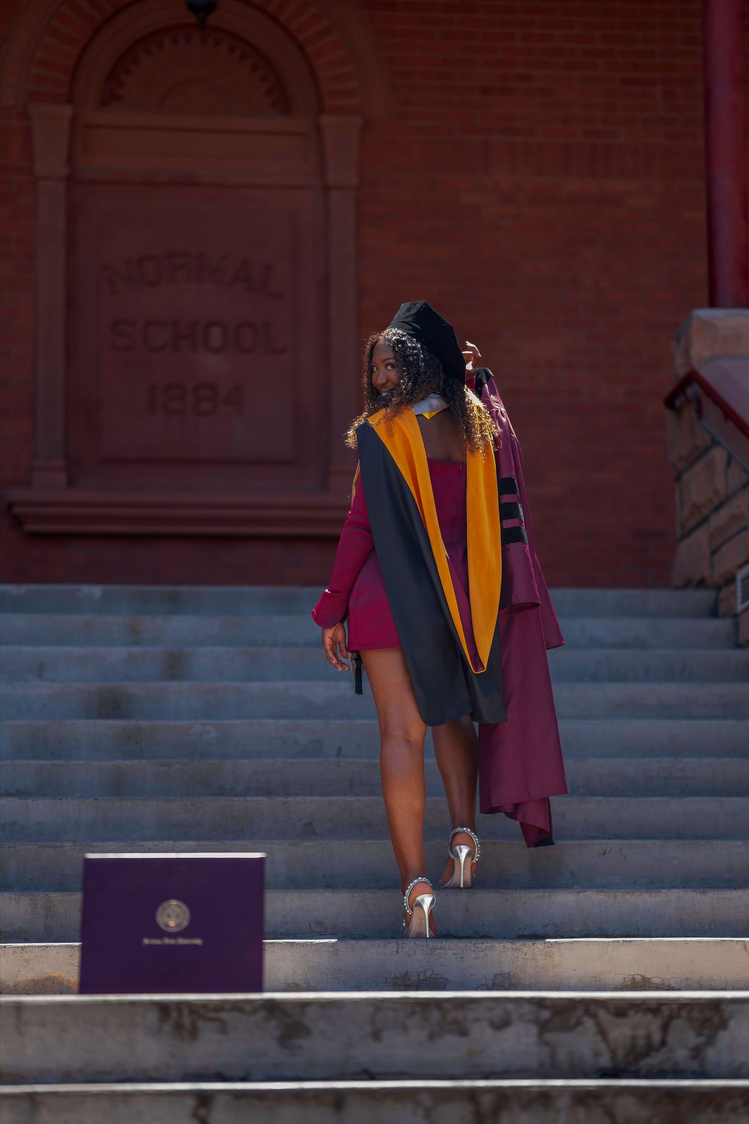 A graduate in maroon gown and cap walks up stone steps outside a historic Normal School 1884 brick building.