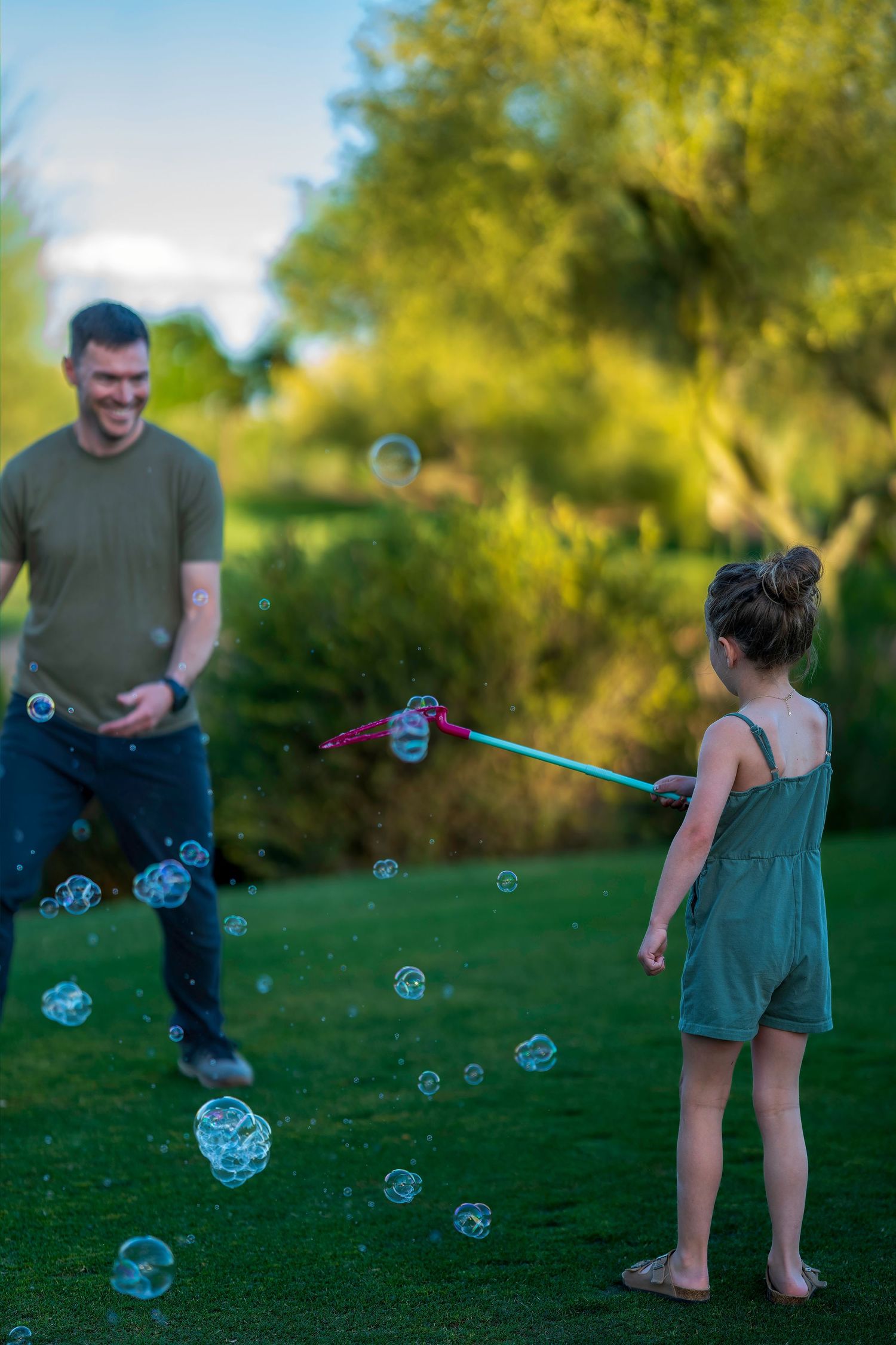 A smiling man watches a young girl blow bubbles with a wand in a lush green backyard.