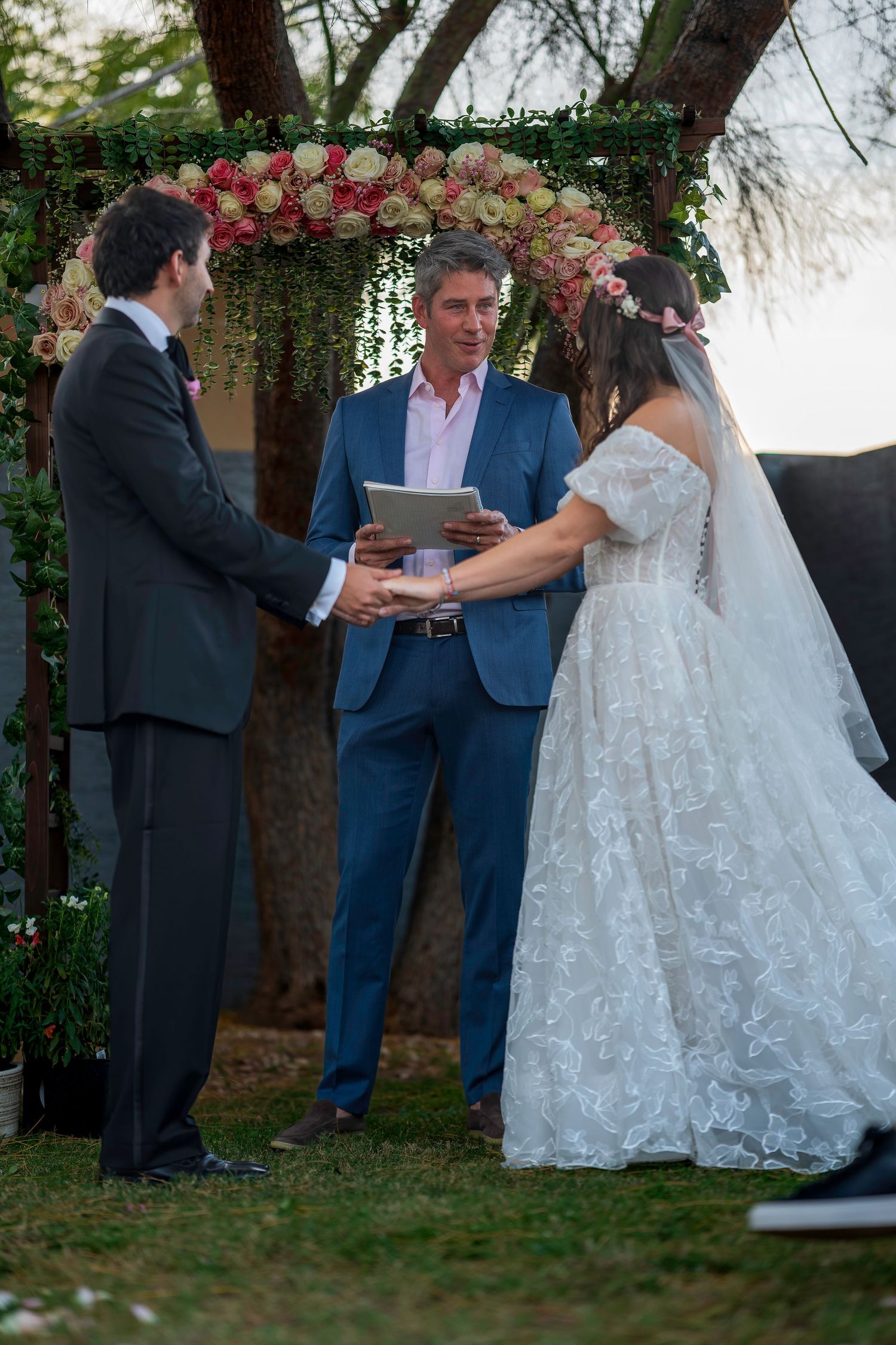 Bride and groom exchange vows outdoors under a floral arch, officiated by a man in a blue suit.