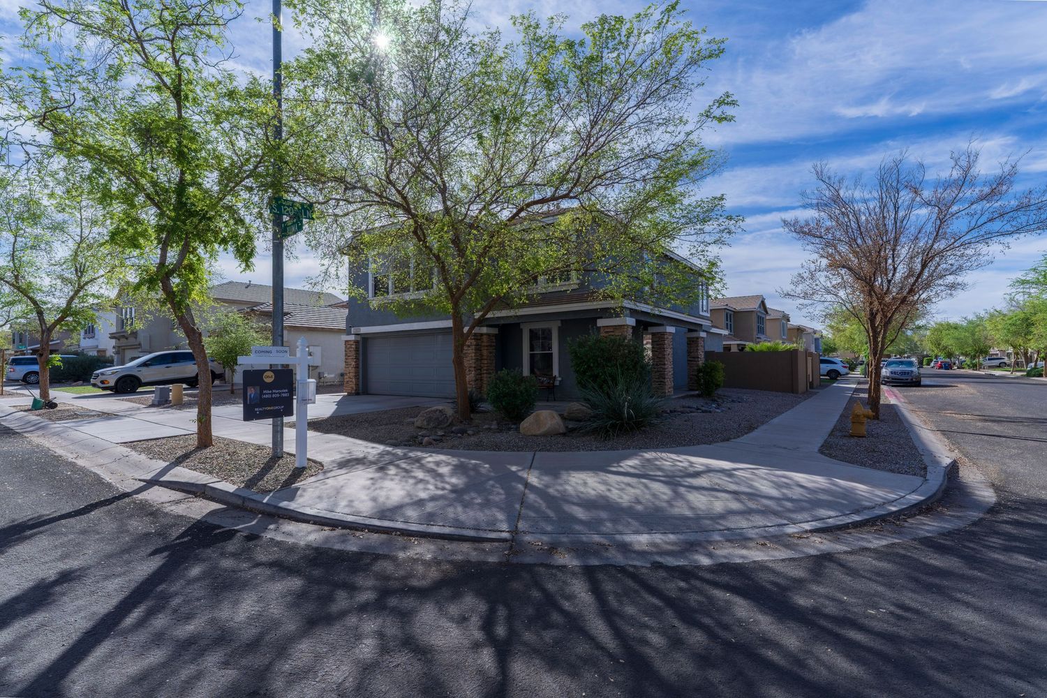 Corner lot home with two-car garage, mature trees, and paved driveway in a sunny Arizona neighborhood.