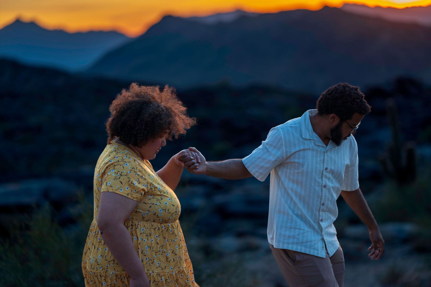 Couple holding hands dancing against a vibrant mountain sunset with colorful orange and purple sky.