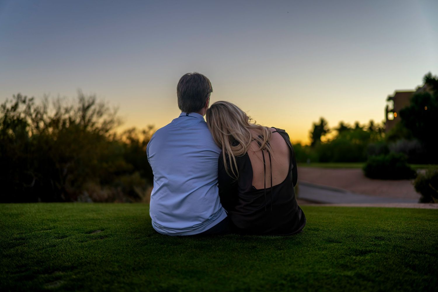 Couple sitting together on grass watching a beautiful sunset with silhouetted trees in background.