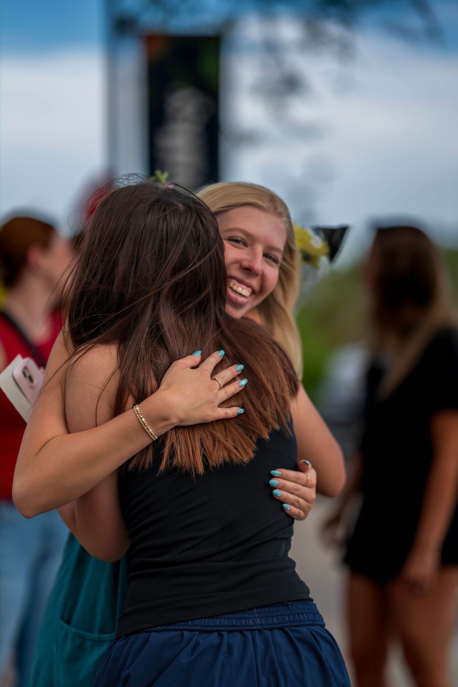 Two young women share a joyful hug outdoors, with one smiling broadly and sporting teal nail polish.