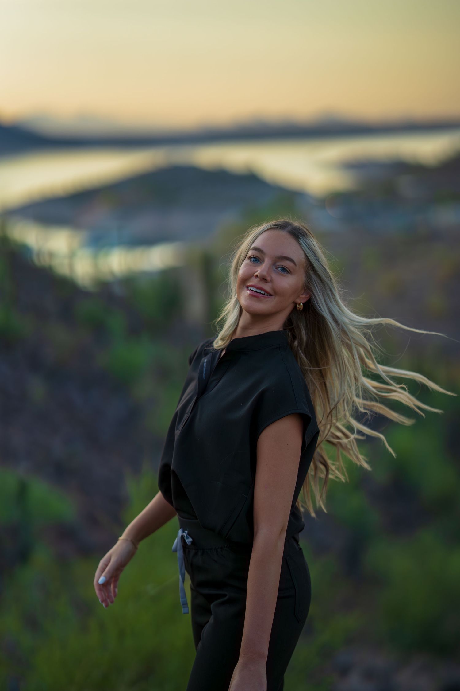 Healthcare worker in black uniform poses with windswept hair against scenic mountain backdrop.