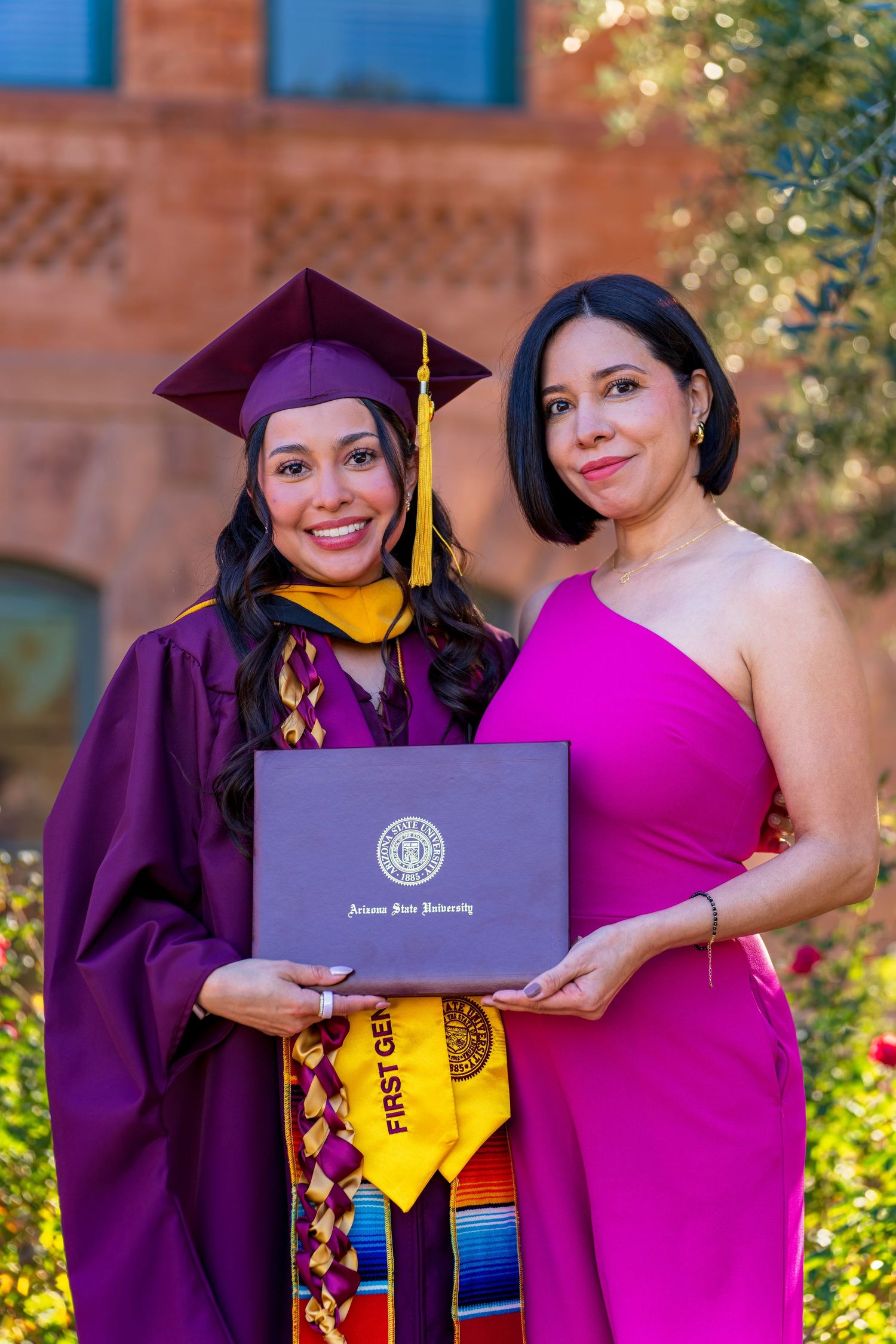 Two graduates in purple regalia holding blue diploma covers while posing in garden setting.