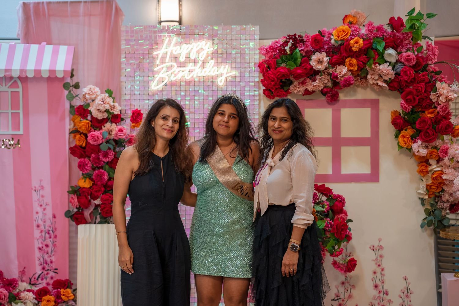 Three people pose in front of a pink floral wall with decorative elements at a celebration.