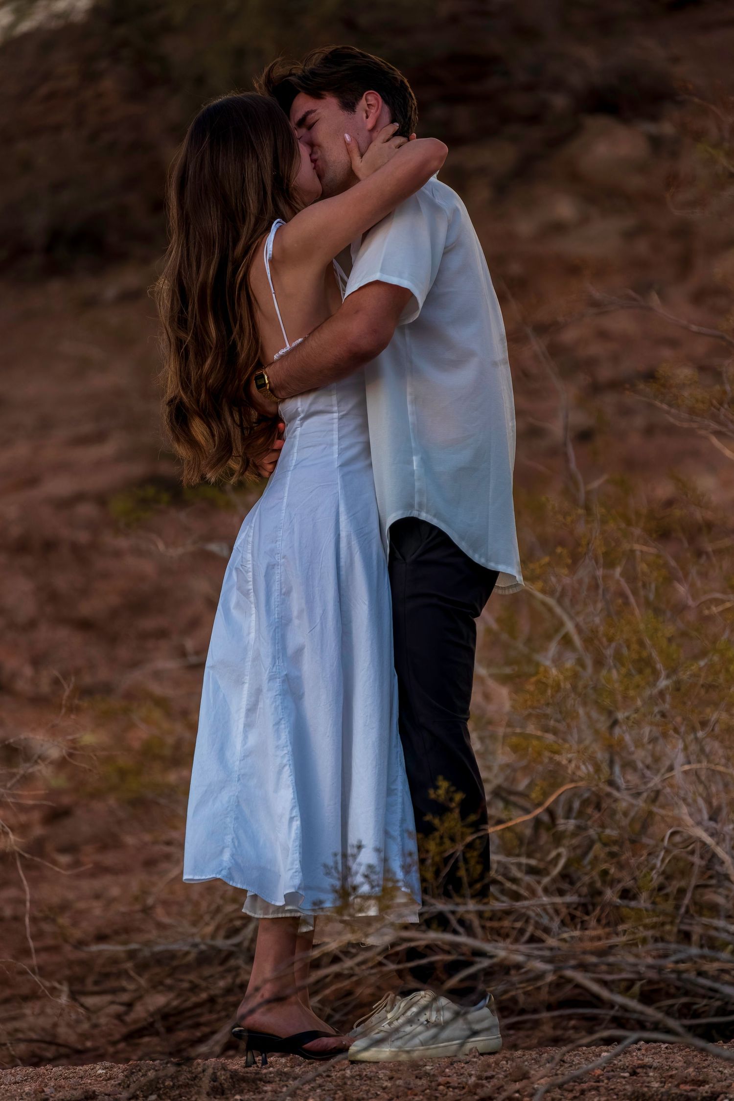 Romantic couple embraces in a white dress and casual attire against a desert landscape at sunset.