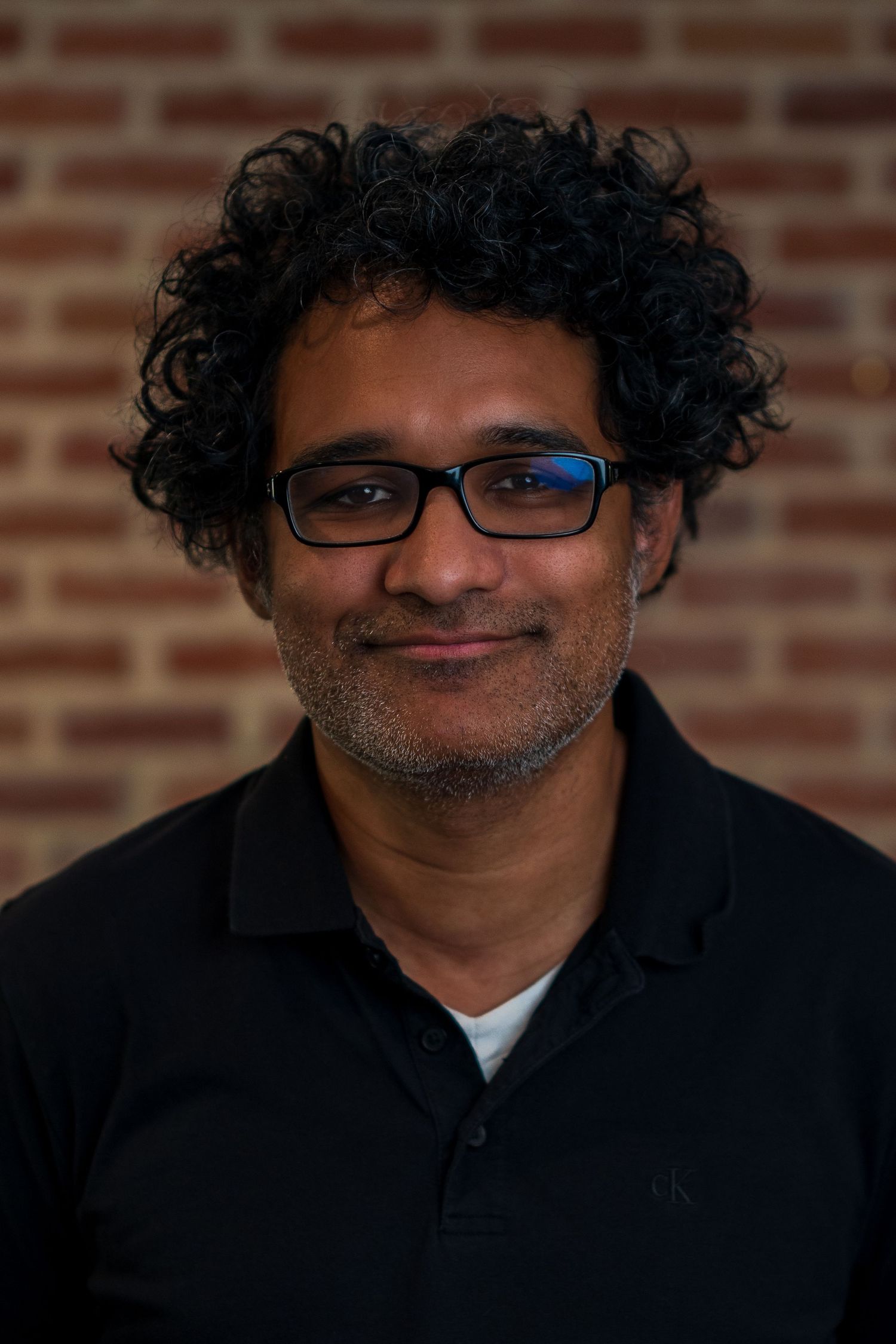 Portrait of a person with curly hair and glasses against a brick wall backdrop.