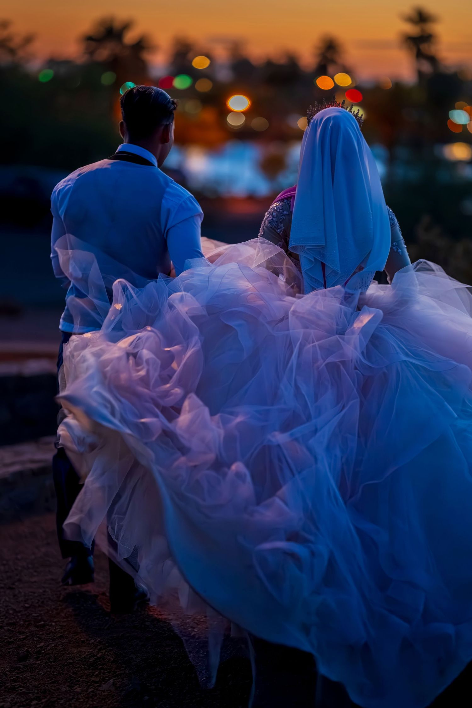 Bride in flowing white gown and veil walks with groom at sunset, city lights bokeh in background.
