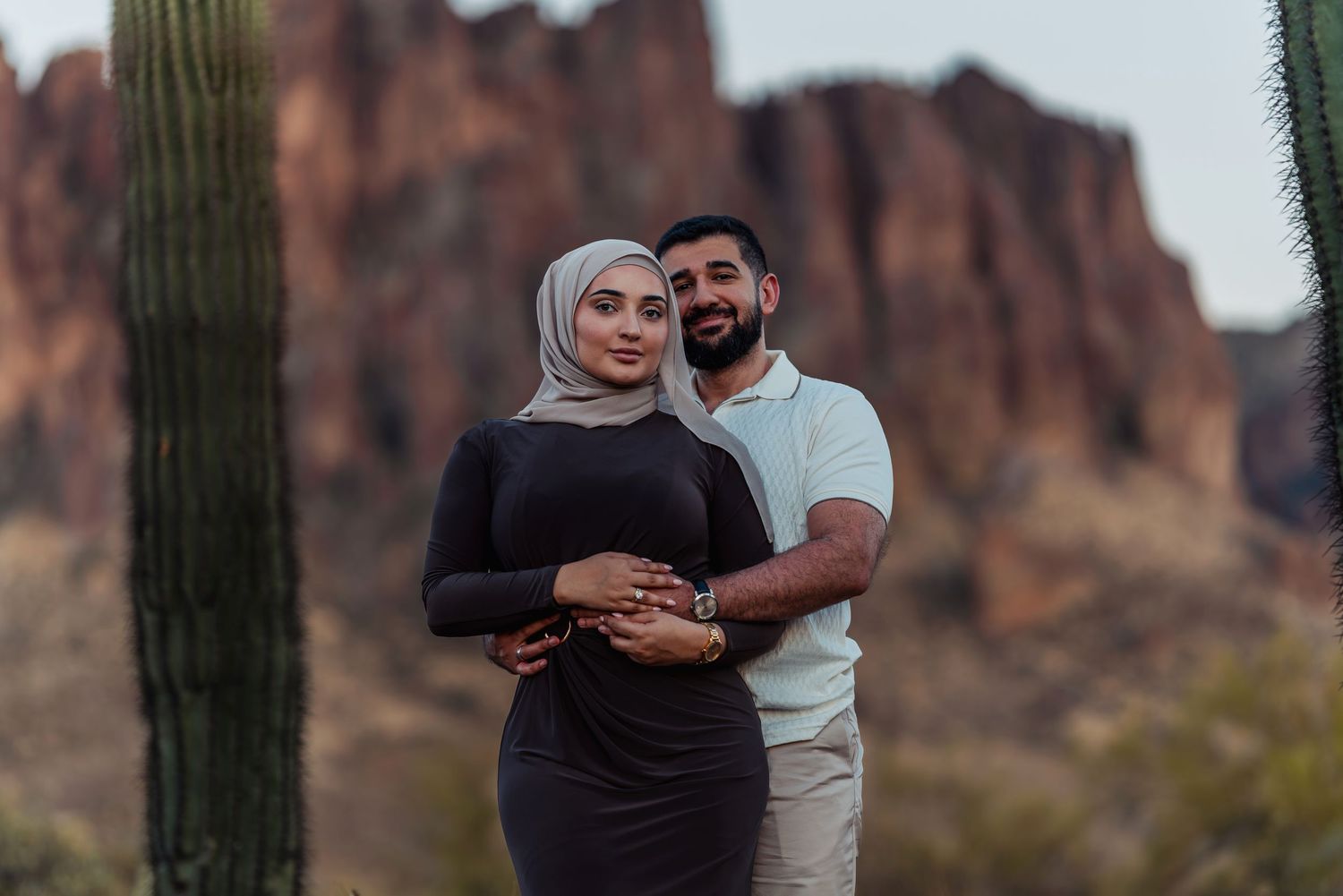 Couple embraces in desert landscape with saguaro cactus and red rock cliffs at dusk.